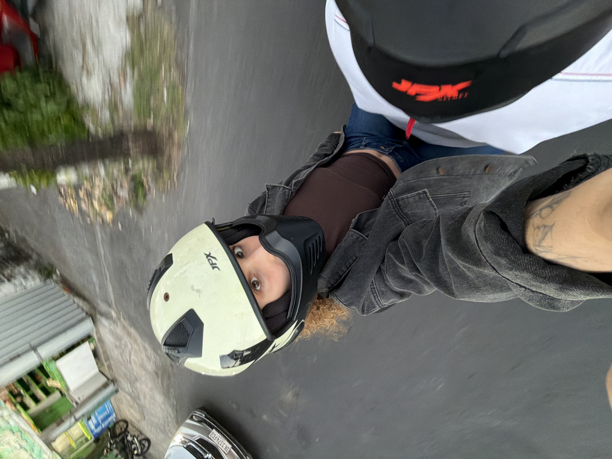An overhead, close-up shot captures a person wearing a cream-colored motorcycle helmet with black accents and a black face mask, looking directly at the camera. Their curly, auburn hair is visible around the helmet. They are wearing a dark brown top and a dark gray denim jacket over blue jeans. Part of a white motorcycle seat with red and blue trim is visible in the lower right corner. The scene appears to be a street in North Kuta, Indonesia, with a silver car visible on the left and a blurred green and white building in the background. A tree and foliage are also present in the background.