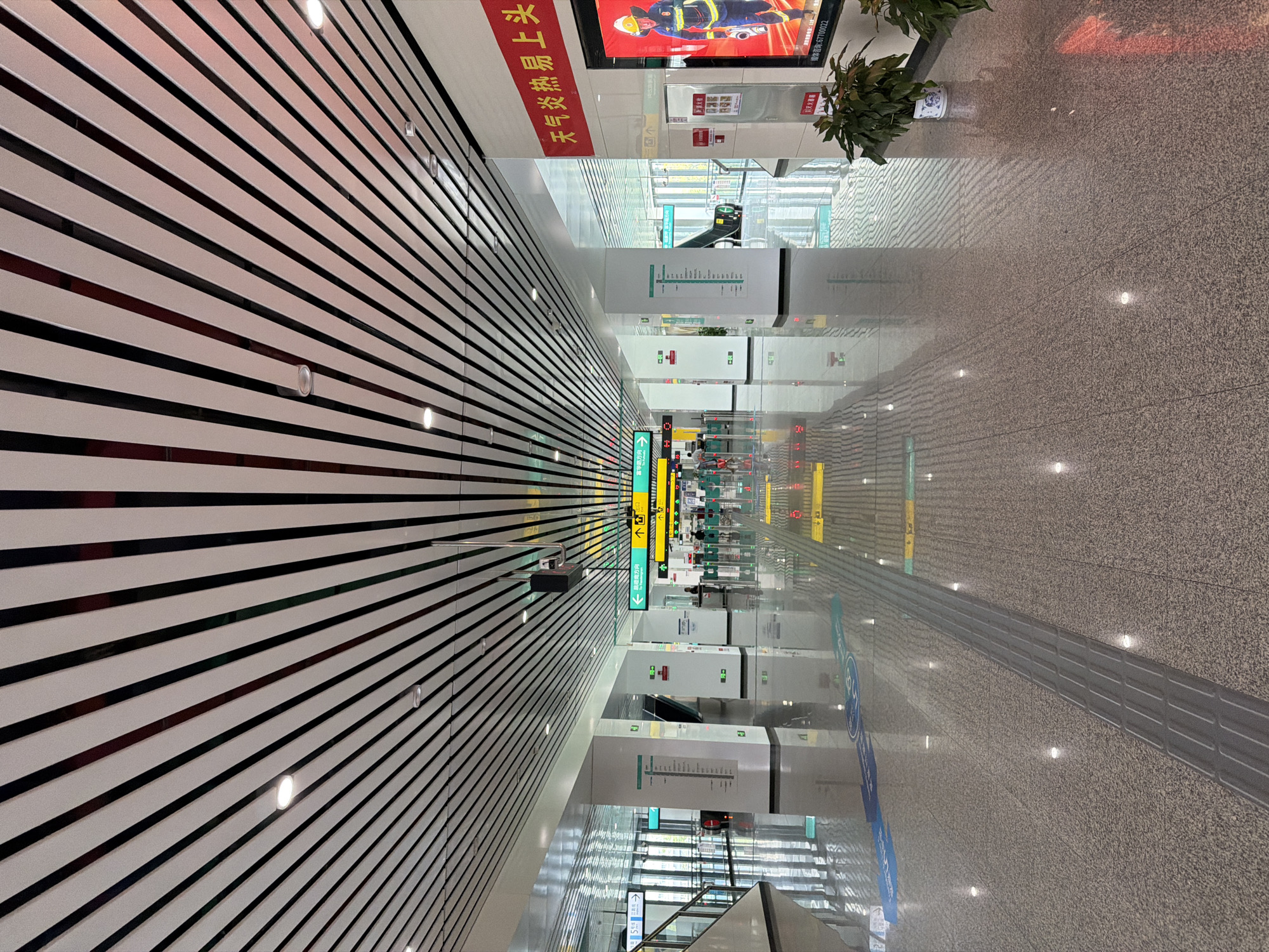 This is a wide-angle, eye-level shot of a modern, well-lit interior space, likely a transportation hub like a subway station or airport terminal, in Tiaodeng, China. The perspective draws the viewer down a long, polished granite floor that reflects the overhead lighting and architectural features.
The ceiling is a dominant element, composed of parallel white slats separated by dark, possibly black, strips. Integrated into the ceiling are circular lights and what appear to be sensors or speakers.
The walls are white and feature large support columns. Various signs with directional arrows and text in Chinese characters are visible, indicating pathways and information. Some signs are illuminated with green light. On the right side of the image, a large red banner with yellow Chinese characters is displayed, and next to it is a digital display showing an image of a person in a yellow and blue uniform holding a tool.
The floor is a speckled granite, with a tactile paving strip running down the center, a common feature for visually impaired individuals in public spaces. Blue and green directional markings are also visible on the floor.
In the background, escalators and glass partitions suggest further areas of the building. A few potted plants are placed strategically, adding a touch of greenery. The overall impression is one of order, cleanliness, and modernity. There are no people clearly identifiable in focus, though some blurred figures can be seen in the distance near the ticket gates.