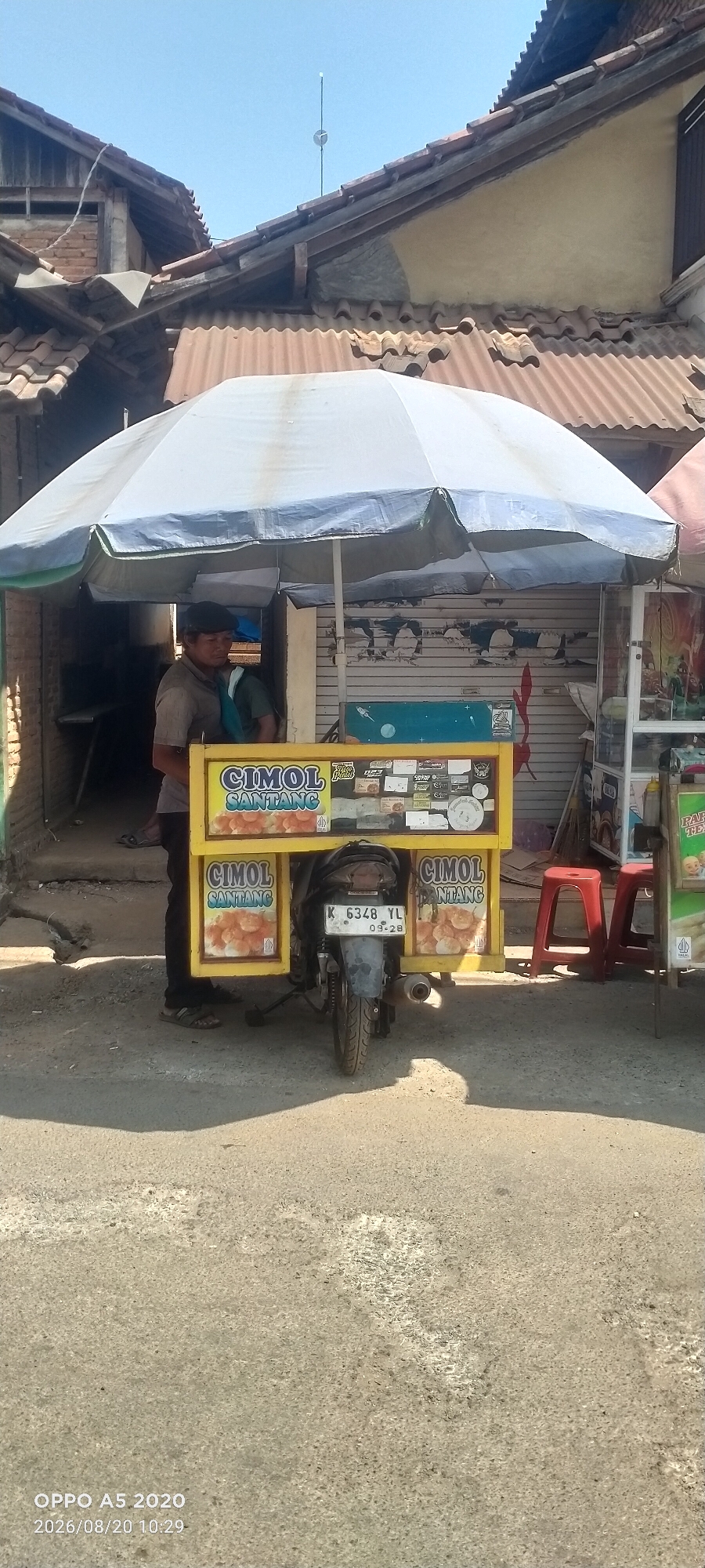 A man stands behind a yellow food cart attached to a motorcycle in Bugel, Indonesia. The cart is covered by a large white umbrella, and its front displays signs that read "CIMOL SANTANG" with images of fried snacks. The motorcycle's license plate reads "K 6348 YL" and the date "09-28". The background shows a building with a corrugated metal roof and a faded shutter. Two red stools are placed next to the cart, and a stack of boxes is visible to the right. The ground is paved and appears to be a street or pathway. The sky is clear and blue. The overall scene depicts a typical street food vendor setup.