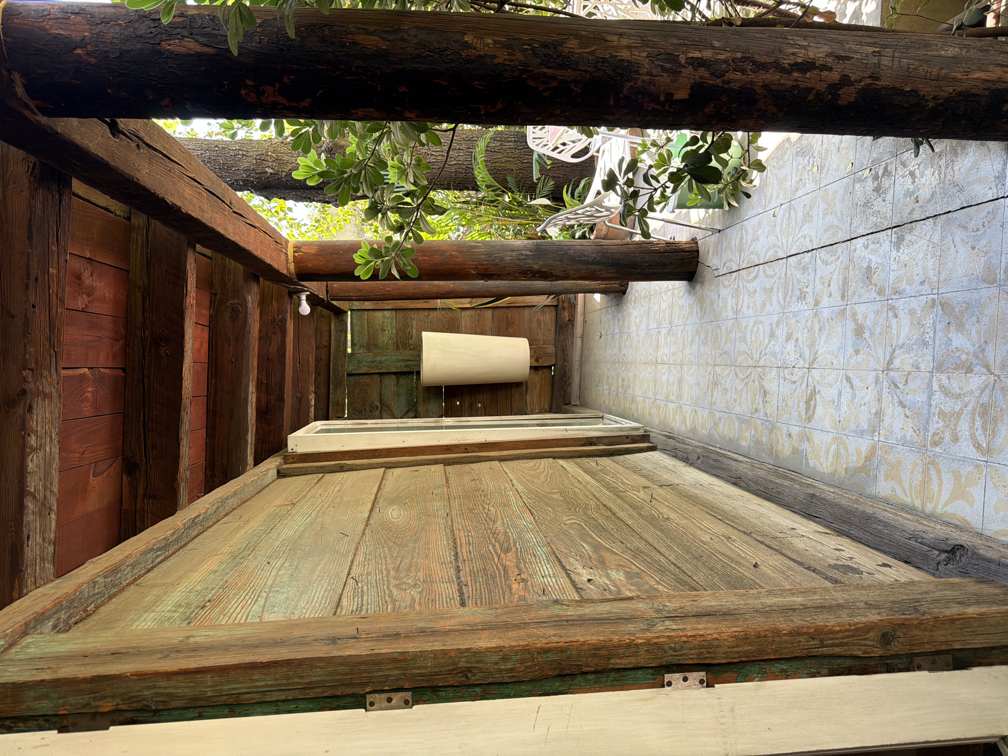The scene depicts a covered outdoor corridor or patio in Puebla, Mexico. A rustic wooden wall, constructed from horizontal planks with weathered green and brown paint, forms the left boundary. A light-colored doorframe with visible hinges is in the immediate foreground on the left. Overhead, a ceiling composed of dark, aged wooden beams and planks is supported by rough-hewn, dark wooden columns, one prominent on the right side. The floor is tiled with a light-colored pattern featuring faded light brown and white/grey ornate designs, and a distinct linear crack extends across its surface. A cylindrical, pale cream-colored wall sconce is mounted on the wooden wall towards the far end of the corridor. A small, white LED bulb is affixed to one of the ceiling beams. Beyond the wooden columns on the right, an outdoor area is visible, partially obscured by green foliage, revealing two white, decorative patio chairs and a portion of a matching table. The background shows large tree trunks and bright natural sunlight, indicating an open exterior environment. No people, actions, or interactions are present. All wooden elements and floor tiles display signs of age and wear.