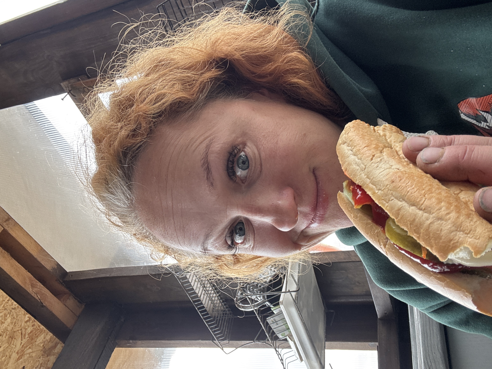 A woman with reddish-blonde, curly hair and light blue eyes looks directly at the camera with a neutral expression, showing a slight hint of a smile. She is holding a hot dog or similar sandwich in her right hand, which appears to be in a baguette-style bun with ketchup and sliced pickles visible. Her hands show some dirt under the fingernails. She is wearing a dark green hoodie, with a graphic design partially visible on her right shoulder.
The image is taken from a low-angle perspective, giving the impression that the woman is looking down at the viewer. The setting is an indoor or covered outdoor space, likely a utility area, shed, or rustic kitchen, characterized by exposed wooden beams and a translucent corrugated roof panel that lets in bright natural light, indicating daytime. In the background, a metal dish drying rack is visible, containing small items. The location context for this scene is Chaikovsky, Russia. There is no visible text in the image.