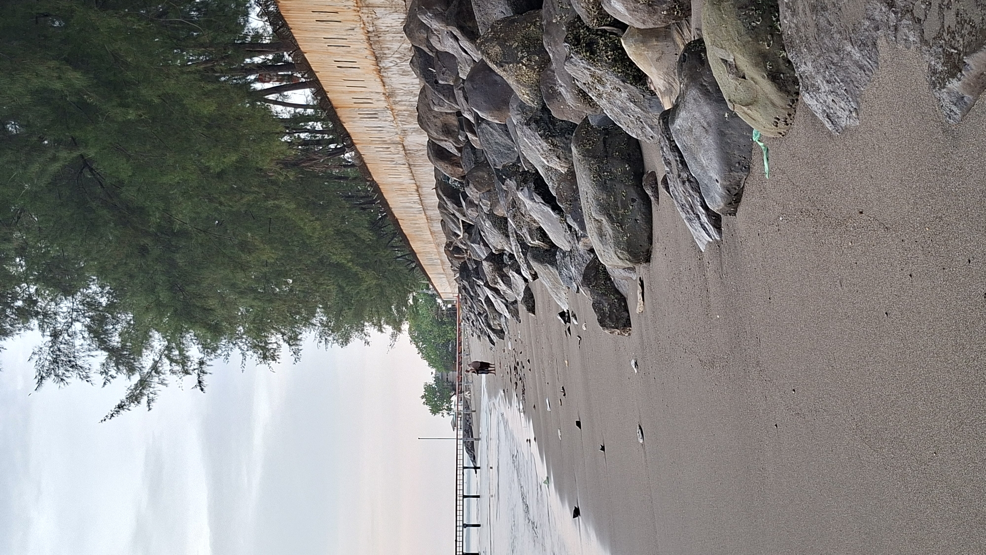 The image shows a beach scene in Lhokseumawe, Indonesia. A sandy beach is visible in the foreground, with the ocean lapping at the shore on the left side. The beach is lined with large rocks, forming a natural barrier, and behind the rocks is a concrete wall topped with trees. In the distance, a pier extends into the water. Two people are walking on the beach, and there is some litter scattered on the sand. The sky is overcast.
