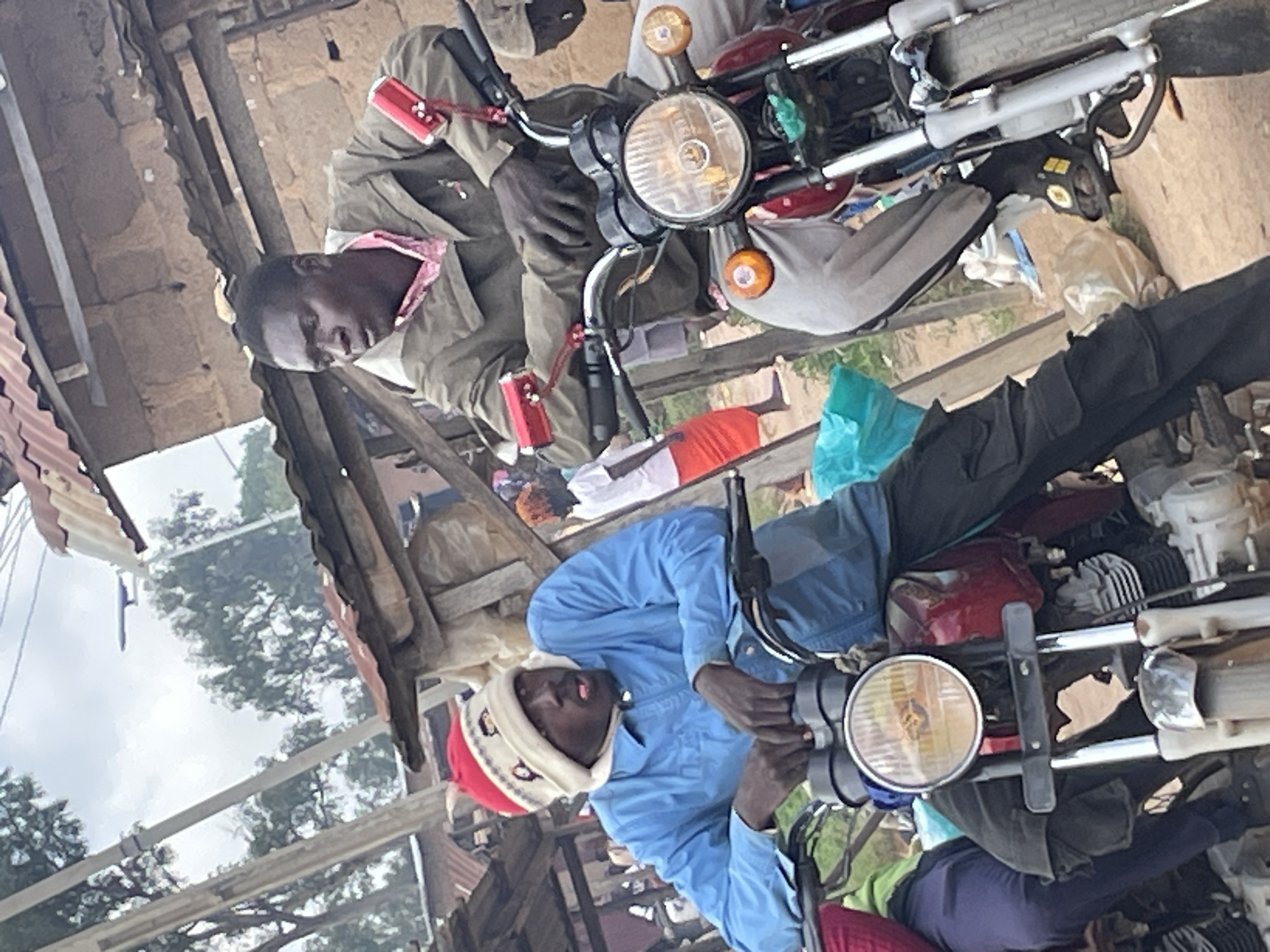 Two people are sitting on separate motorcycles, seemingly preparing to depart or having just arrived. The image is taken outdoors in what appears to be a rural or market setting in Wang, Nigeria. The lighting suggests it is daytime, possibly late morning or early afternoon, with diffused light indicating a cloudy or overcast sky.
In the foreground, a person wearing a light blue jacket and a distinctive red and white patterned beanie is seated on a motorcycle. They have their eyes closed, and their mouth is slightly open, giving the impression of rest or weariness. The motorcycle in the foreground is red.
Behind this person and slightly to the left, another person is seated on a separate motorcycle. This individual is wearing a green jacket over a pink garment and is looking down. The motorcycle is a darker color, possibly red or maroon.
Both motorcycles are parked close together, with various items, including bags and what looks like folded fabrics, piled around them. The background shows a rustic structure made of wood and possibly corrugated metal, typical of informal market stalls or housing in the region. Some trees are visible through the gaps in the structures, and the overall impression is one of a busy, functional outdoor space. There is no visible text in the image. The scene feels relaxed but functional, suggesting a moment of pause in daily activities.