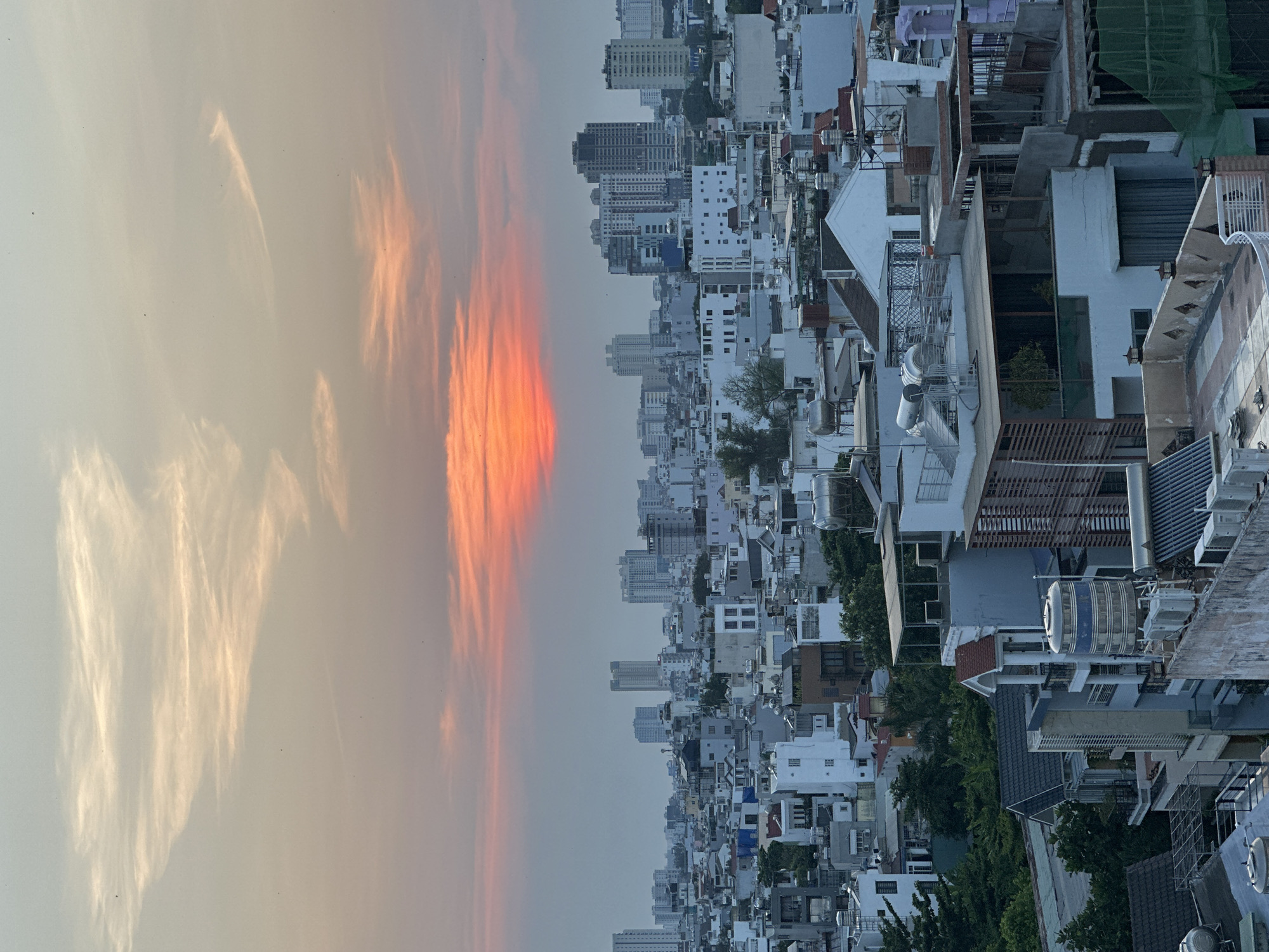 The image displays a sprawling urban landscape of Ho Chi Minh City, Vietnam, captured at either sunrise or sunset, viewed from a high vantage point. The sky occupies the upper half of the frame, showcasing a dramatic transition of colors. Vibrant streaks of orange and deep yellow clouds, illuminated by the low sun, dominate the left and center, suggesting the "golden hour." These intense hues fade into softer, lighter yellow and gray tones towards the right, with delicate, wispy cloud formations scattered across the expanse.
Below the captivating sky, the city stretches out densely. A vast array of buildings, varying in height and design, fills the lower half of the image. The foreground and midground are characterized by numerous white and light-colored structures, many with flat or gently sloped roofs, exhibiting typical Vietnamese urban architecture. Rooftops are replete with functional elements such as cylindrical water tanks, air conditioning units, and various utility structures. Some taller, more modern high-rise buildings are visible in the hazy distance, marking the city's evolving skyline. Small patches of green foliage and trees are interspersed among the concrete, particularly on or between buildings.
The absence of people highlights the scale of the urban environment and the quiet, contemplative atmosphere of the moment. The soft, diffused light from the twilight hours casts a serene glow over the entire scene. There is no visible text in the image.