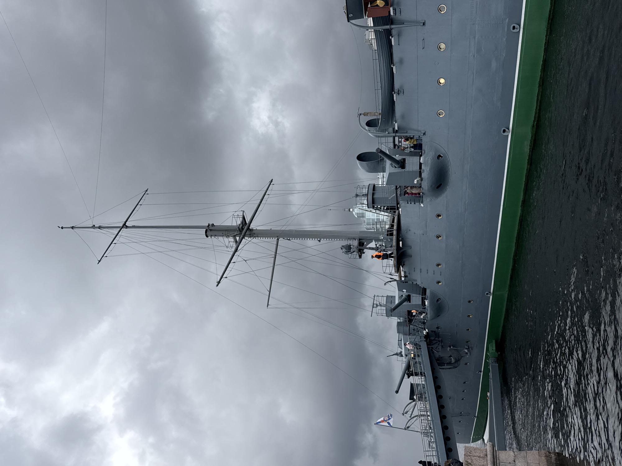 The image shows the cruiser Aurora, a museum ship and a landmark in Saint Petersburg, Russia. The ship is docked, with its port side facing the viewer. The hull is painted gray, with a green stripe along the waterline. A tall mast rises from the deck, supported by numerous wires. Several cannons are mounted on the deck. People, some wearing orange safety vests, are visible on the deck. A flag with a white field and a blue cross is displayed at the stern. The sky is overcast with dark, ominous clouds. Water is visible next to the ship, indicating the Neva River or a canal. The background suggests a city environment, though details are obscured by the weather.