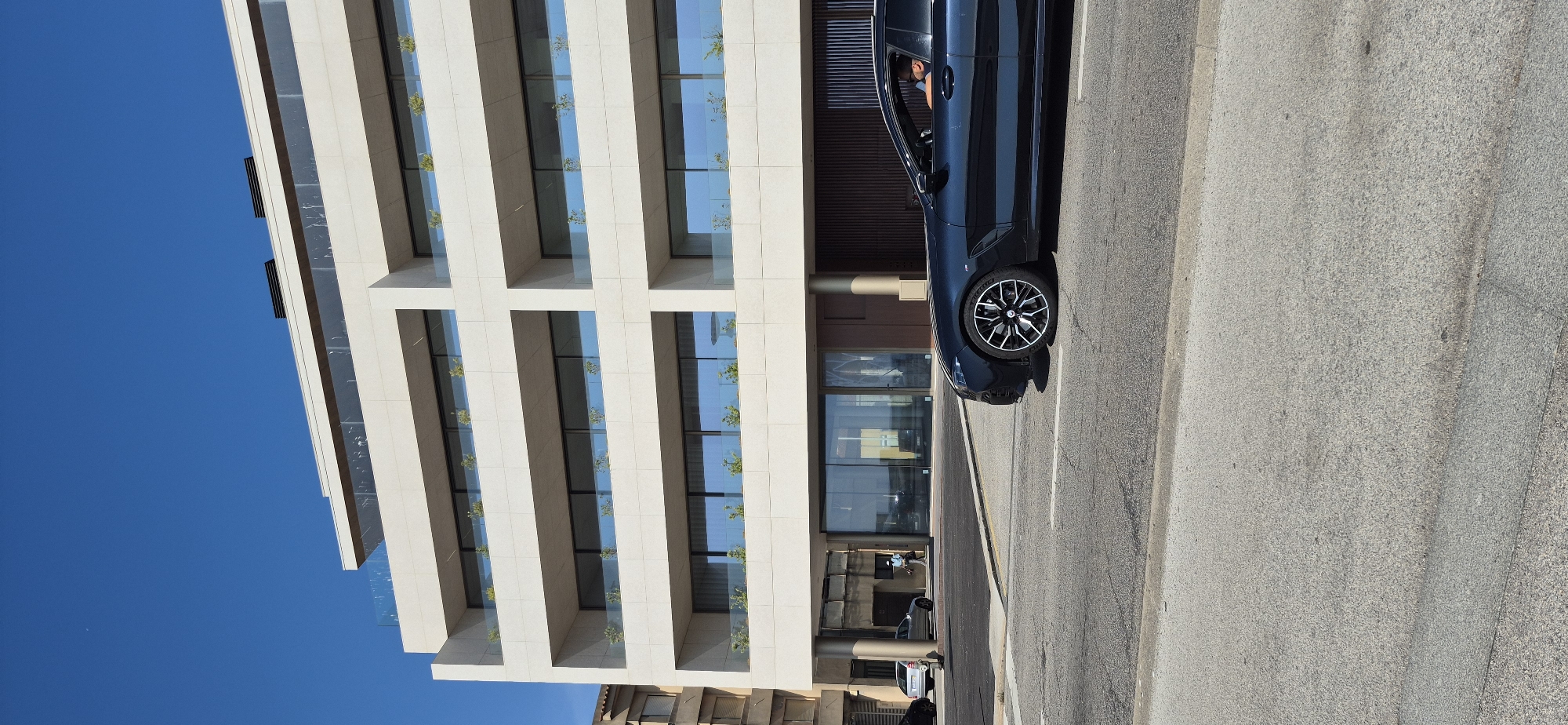 A navy blue car with a black interior and tinted windows is driving on a street in Vila do Conde, Portugal. The car is in the foreground, and its front passenger side is visible. The driver, a white male with dark hair and a beard, is wearing a black t-shirt and sunglasses. He is looking forward. The car has a black roof and a black trim around the windows. The wheels are black with silver spokes. Behind the car, there is a modern building with multiple levels. The building has a cream-colored facade with rows of balconies and large windows. Small plants are visible on the balconies. The sky is clear and blue. To the left of the building, another building with a darker facade and brown window frames is partially visible. In the background, there are two silver cars parked on the side of the street and a person walking. The street is paved with asphalt and has white lane markings.