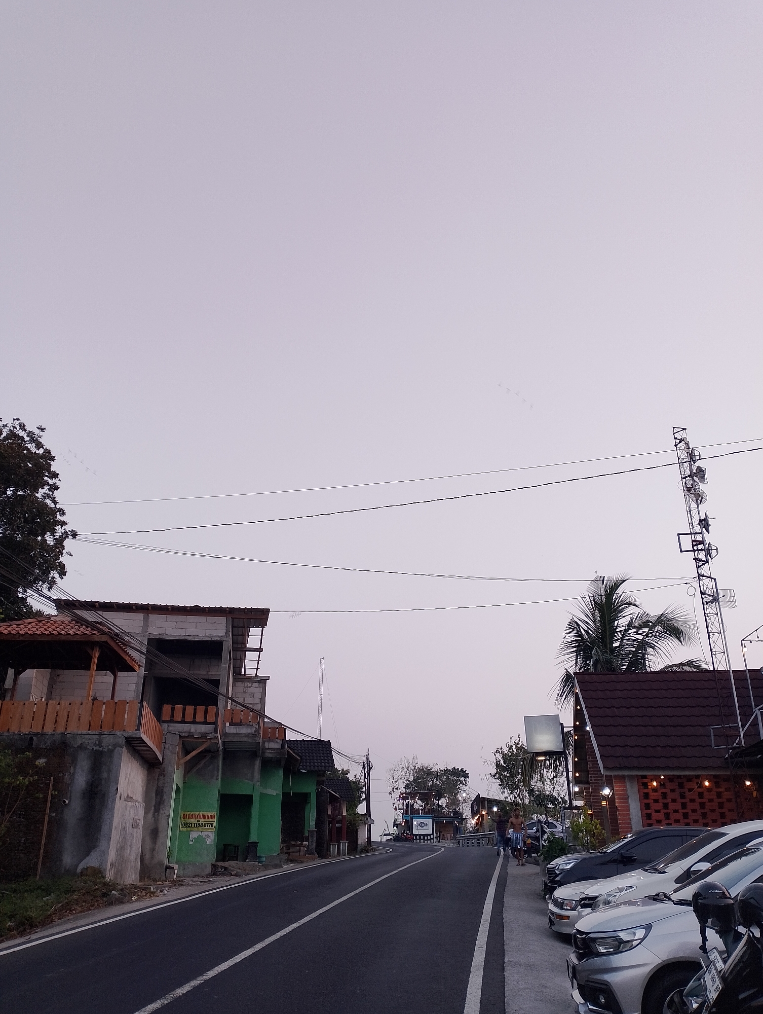 The image captures a street scene in Srimulyo, Indonesia, under an overcast sky. On the left side of the asphalt road, a multi-story building is under construction, with exposed concrete blocks and wooden scaffolding. A small wooden structure with a red tiled roof is attached to the lower level. A green sign with white lettering is affixed to the concrete wall of the building.

Further down the road, on the right, a building with a dark red tiled roof and brick accents features outdoor seating illuminated by string lights. Several parked cars, including a silver Honda, a white car, and a black car, are lined up along the sidewalk in front of this establishment. Two motorcycles are also parked in the foreground.

In the background, a telecommunications tower stands tall. A few people are visible walking along the sidewalk near the restaurant. The overall impression is that of a developing or revitalized area along a main road, possibly a commercial district or a popular spot for dining and socializing.