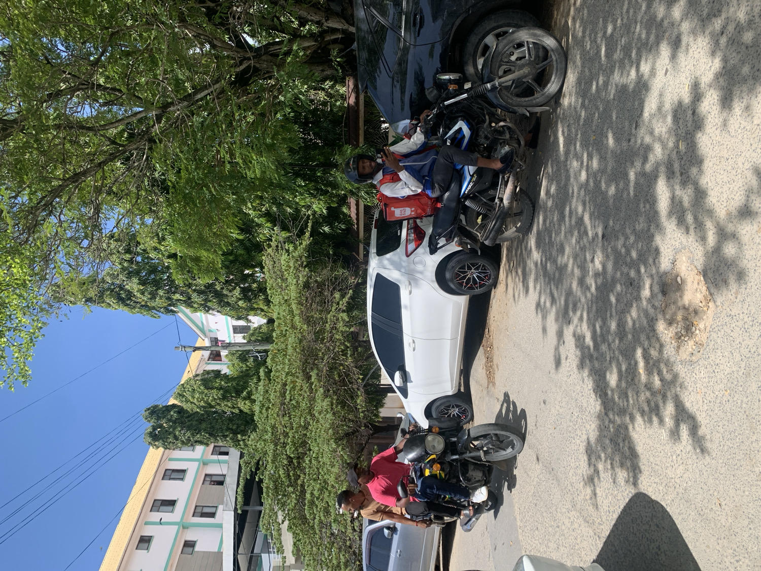 The image depicts a sunlit urban street scene in Dar es Salaam, Tanzania, featuring people, motorcycles, and cars parked alongside a road lined with trees and buildings.
Several men are present. In the mid-ground, one man wearing a red and white jacket, characteristic of a delivery rider, is seated on a black and blue motorcycle, which has a red delivery box on its rear. Another man is standing near this motorcycle. In the foreground, partially visible on the left, a third man in a red shirt is standing next to a black motorcycle.
Multiple vehicles are parked along the side of the street. A white car is visible in the mid-ground, parked in front of a black car. The white car features black wheels with distinctive red accents. A silver or grey car is partially visible in the foreground, to the left of the man in the red shirt. At least three motorcycles are visible, positioned near the parked cars.
The setting includes a vibrant natural environment with numerous green trees and bushes casting dappled shadows across the asphalt road. In the background, several multi-story buildings with light-colored facades and some with yellow/orange roofs are visible under a clear blue sky. A utility pole with power lines stands against the sky.
The activity appears to be people gathered or waiting by their vehicles, possibly taking a break or conversing, suggesting a typical daily street scene or a stop for delivery personnel. The bright sunlight and distinct shadows indicate it is daytime, likely mid-morning or afternoon, with fair weather. No discernible text is visible in the image.