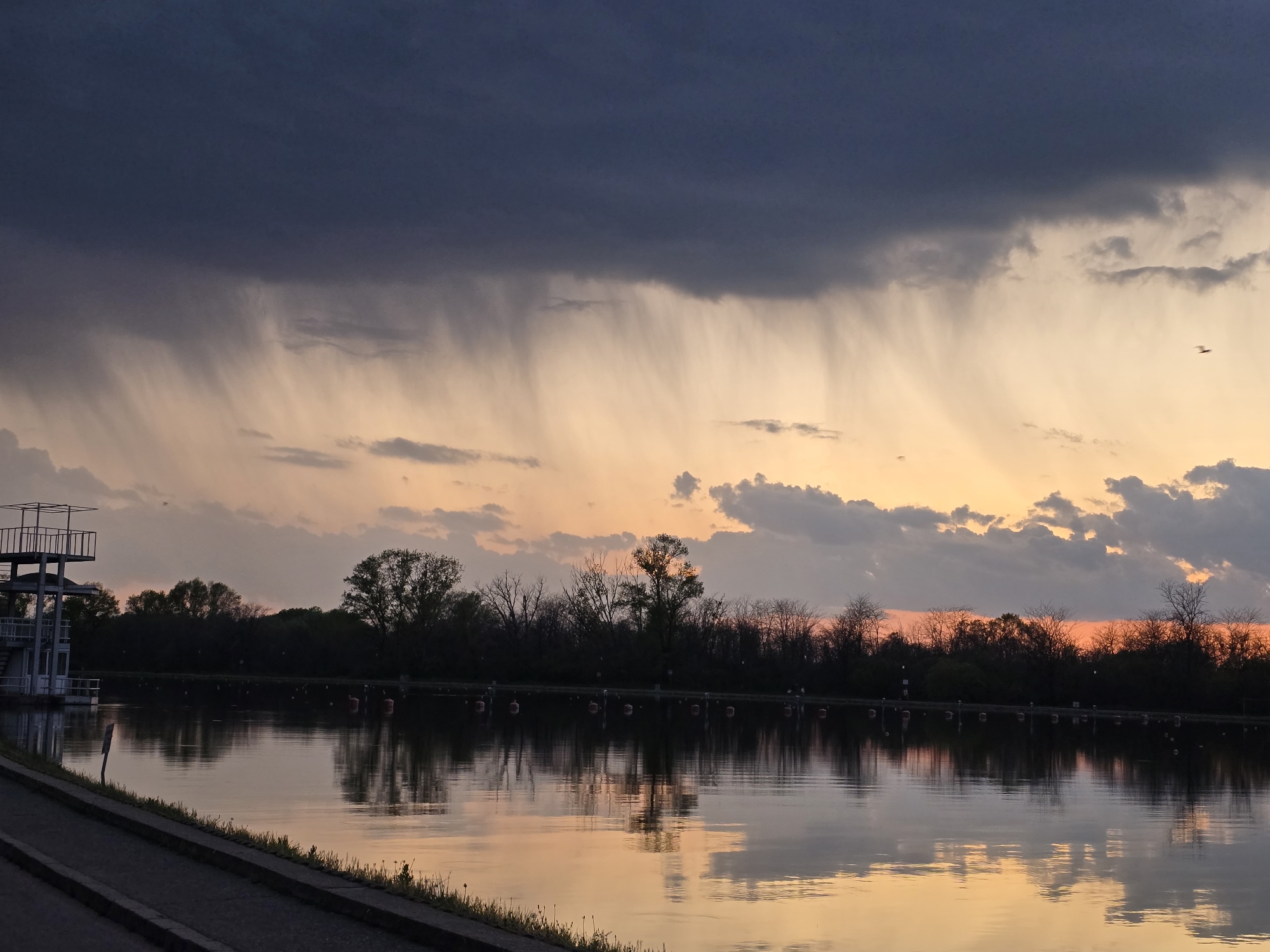 A wide shot captures a body of water with a shoreline, trees, and a building on the left under a dramatic sky. The sky transitions from a dark, ominous storm cloud at the top to a lighter, orange-tinted sunset glow near the horizon. Streaks of falling rain are visible beneath the dark cloud. The water reflects the colors of the sky. A single bird is visible in the sky. A line of trees is silhouetted against the sunset. On the left, a multi-level structure with metal scaffolding extends from the water. The shoreline features a concrete edge. The location is Unknown, Unknown.