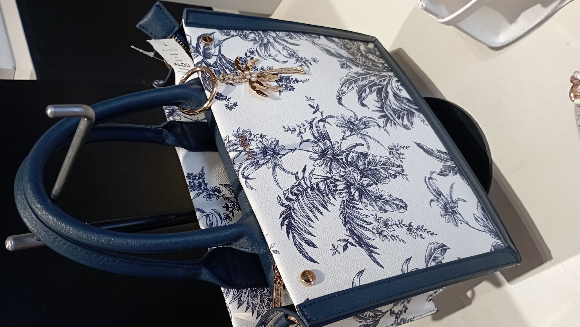 A close-up shot shows a white Aldo handbag with a blue floral pattern. The bag features blue trim and handles, and a gold-colored palm tree charm is attached. It sits on a surface that transitions from black to white. The scene is likely indoors.