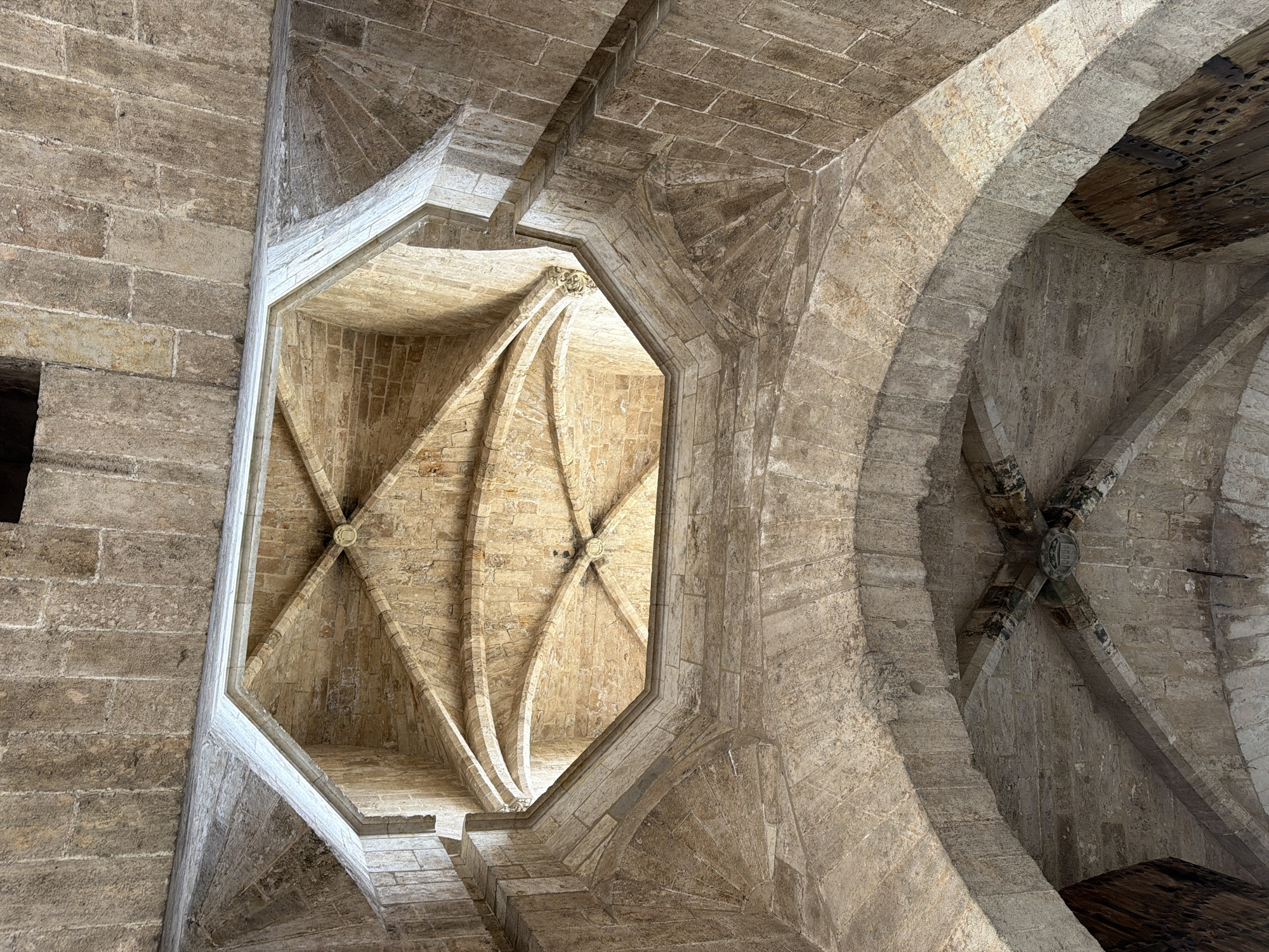 An upward shot of a stone vaulted ceiling. The ceiling is constructed from many large, rectangular stone blocks. There are multiple archways and ribbed vaults, creating a complex geometric pattern. In the center of the image, there is an octagonal opening with a vaulted ceiling above it, allowing light to enter. The style appears to be Gothic or Romanesque architecture.