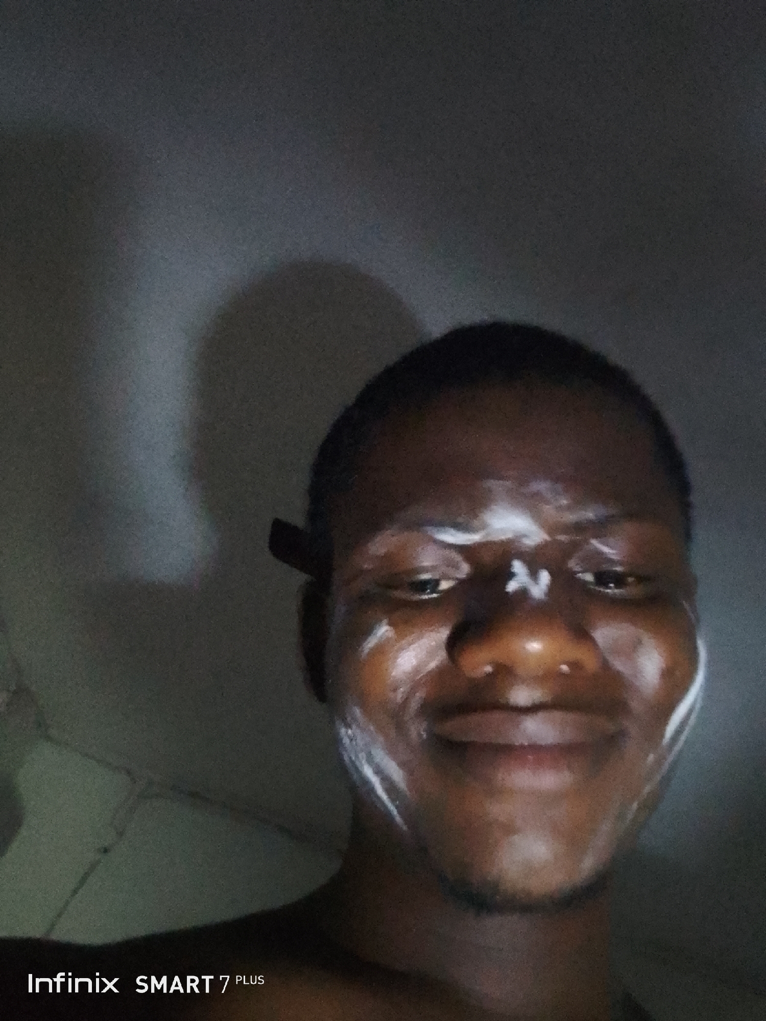 A low-light, close-up portrait shows a person with dark skin looking directly at the camera with a subtle smile. The person's face is partially covered with white cream or a similar substance, appearing as streaks and dots on their forehead, around their eyes, on their nose, and along their jawline. A distinct "N" or "Z" shape is visible in cream on the bridge of their nose, and a curved streak across the forehead.
The background is dark and appears to be an indoor wall, with vague shadows suggesting the presence of other objects or the person's own shadow. The lighting is dim and casts soft highlights on the cream and the person's skin, giving a slightly glossy appearance. Behind the person's right ear, a dark, thin object, possibly a comb or a part of headphones, is visible. The person has short, dark hair.
In the bottom left corner of the image, the text "Infinix SMART 7 PLUS" is clearly visible, indicating the device used to capture the photograph. The overall impression is of a casual, personal photo, possibly taken during a skincare routine or a similar activity.