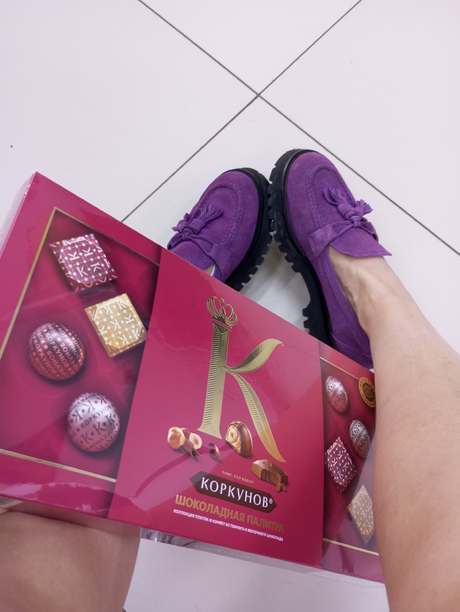 The image is a low-angle, close-up shot of a person holding a box of Korkunov chocolates in front of their legs, wearing purple shoes. The background consists of white floor tiles.
The chocolate box is red and features a large gold "K" logo with a crown on top, along with an image of hazelnuts and chocolate candies. Text on the box reads "Korkunov" and "Shokoladnaya Palitra" (Chocolate Palette) in Russian. The box also displays various types of Korkunov chocolates in individual compartments.
The person in the photo is wearing purple suede shoes with black soles and tassel details. Their legs are visible behind the chocolate box.
The photograph was not taken in the city of Izhevsk, Russia.
