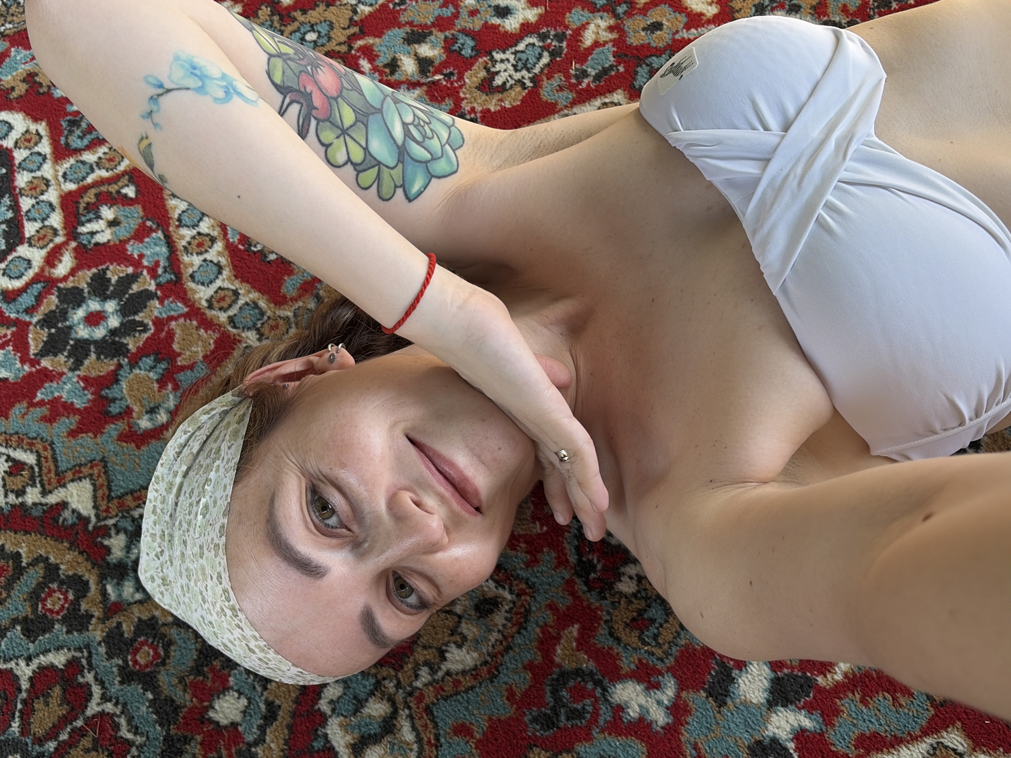 A fair-skinned woman with light brown hair and hazel eyes lies on a patterned rug. She wears a white headband and a strapless white top. Her left arm is raised, showing tattoos on her upper arm. A red string bracelet is visible on her wrist, and a ring on her finger. The rug has a detailed, colorful pattern with reds, blues, browns, and greens. The location is not specified in the image.