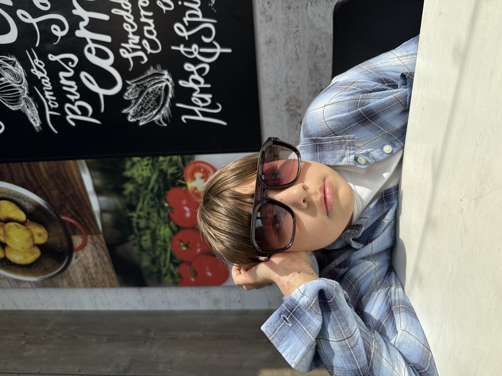 A child with brown hair is wearing sunglasses and a blue plaid shirt while sitting next to a sign. The sign contains a menu of food items, including "Tomato, Buns, Corn, Shredded Carrots, Herbs & Spices," accompanied by illustrations of food items. The child is resting his head on his hand. The location is unknown and is not identifiable as Шебунино, Russia, from the details provided.