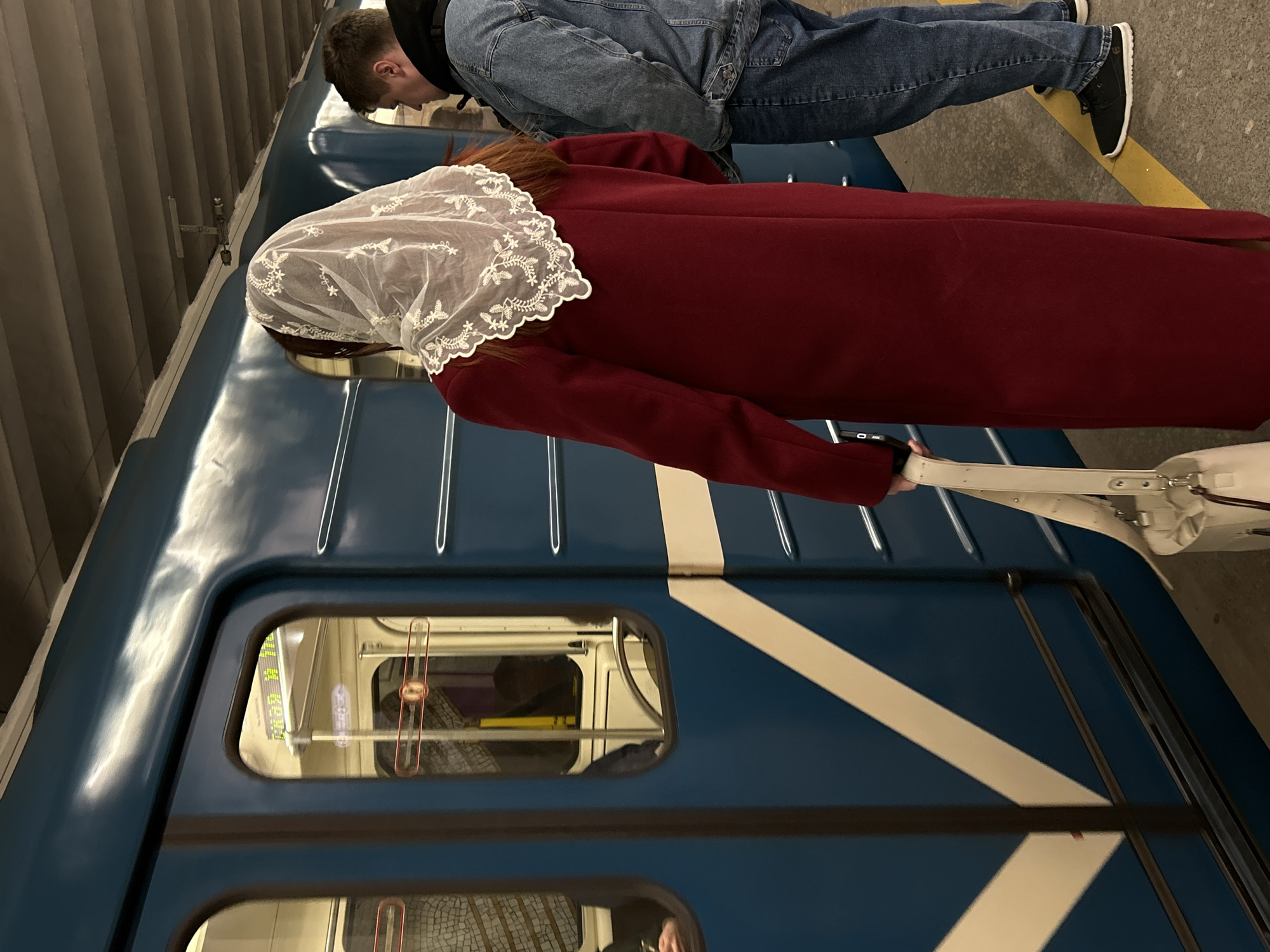 The scene captures an interior or exterior view of the Saint Petersburg Metro, Russia, evidenced by the distinctive blue and white angled stripe exterior of the train car and the design of the train cars, which is typical of the Saint Petersburg Metro.
Two individuals are visible: a person wearing a red coat and a white lace head covering stands near the train car, holding what appears to be a white handbag, and another person wearing jeans and a black top is positioned above near the train car's roof.
The photograph is taken on the platform level of a station, with a segment of the platform and the subway train visible.