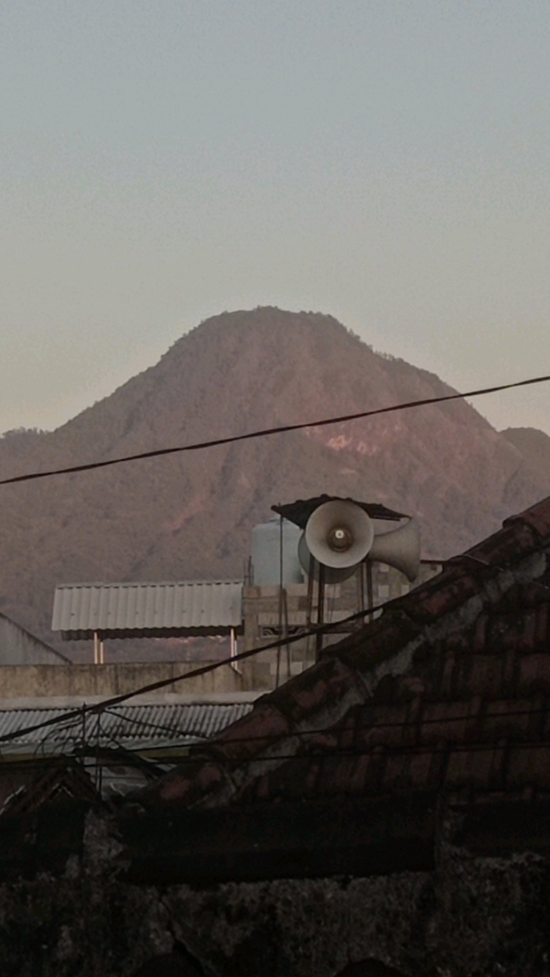 A wide shot, captured at an outdoor setting, shows a mountain in the background, its slopes covered in vegetation and appearing somewhat arid. In the foreground, several structures are visible, including a tiled roof in the lower right corner, a corrugated metal roof in the middle ground, and a large cylindrical water tank. Two public address speakers, mounted on poles, are positioned on top of a building in the middle distance. The sky above is clear and shows a pale, hazy gradient. The overall lighting suggests either dawn or dusk. The image has a rustic, somewhat neglected feel, with exposed wires crisscrossing the frame. The location of Mulyoagung, Indonesia is suggested by the prompt.