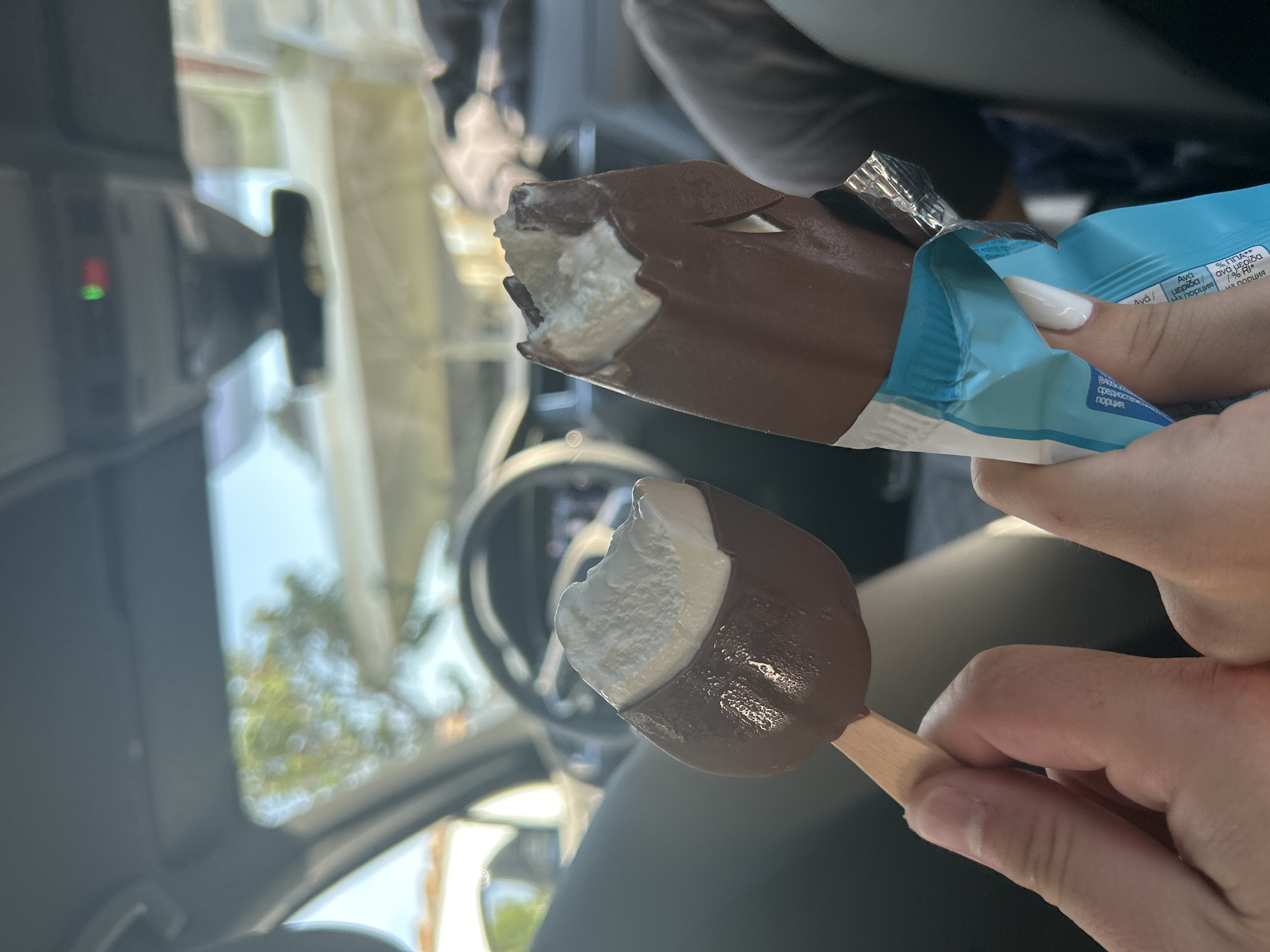 Two partially consumed chocolate-dipped ice cream bars are held by two distinct hands inside a car in Fanari, Greece. The hand on the left holds an oval-shaped ice cream on a wooden stick, with a bite taken from the white interior. The hand on the right, featuring a white manicured fingernail, holds a larger, rectangular chocolate-dipped ice cream, partially unwrapped from a blue and white foil package, also with a bite taken. The blurred background reveals the interior of a modern vehicle, including a steering wheel, dashboard, a rearview mirror, and an overhead console with a green illuminated light. Through the car windows, a bright blue sky and out-of-focus green foliage are visible, indicating an outdoor, daytime environment.