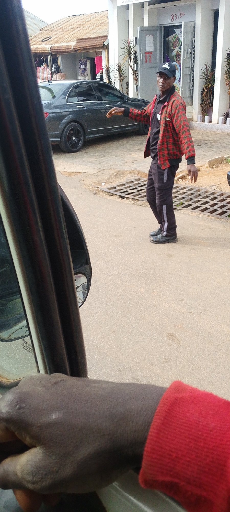 A person wearing a red and black plaid jacket, black pants with white stripes, a black baseball cap, and black shoes stands on a paved road in front of a building. The person's left arm is extended, palm facing upwards, in a gesture of stopping or signaling. Behind the person, a dark-colored sedan is parked. The building has a white facade with multiple glass doors, and signage indicating "83/84." The ground is a mix of asphalt and concrete with a metal grate covering a drain. The scene appears to be a street in Jos, Nigeria, with elements of urban infrastructure and commercial activity.