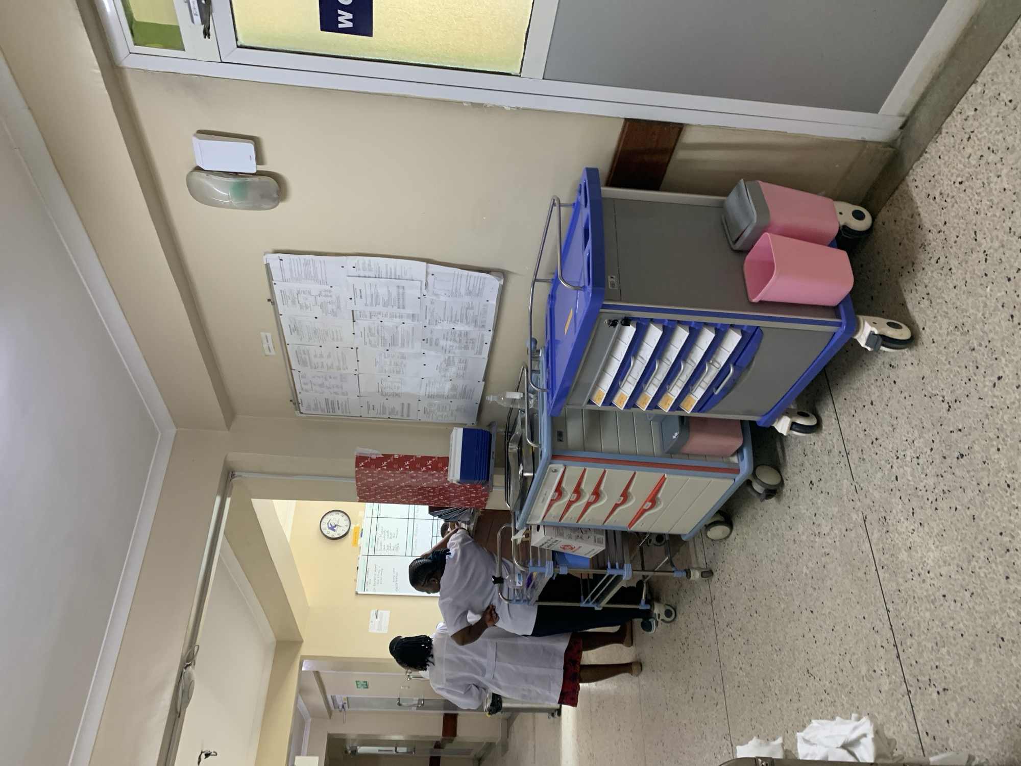 The image shows a hospital corridor. Two individuals wearing white coats are standing near a medical cart laden with supplies, possibly interacting with it. The cart has multiple drawers and storage compartments, with some items visible. The corridor walls are painted in light colors, and a bulletin board with various documents is affixed to one wall. A clock is visible further down the corridor. The scene appears to be indoors, specifically within a medical facility. Without additional information, it's impossible to verify the location within Dar es-Salaam, Tanzania.