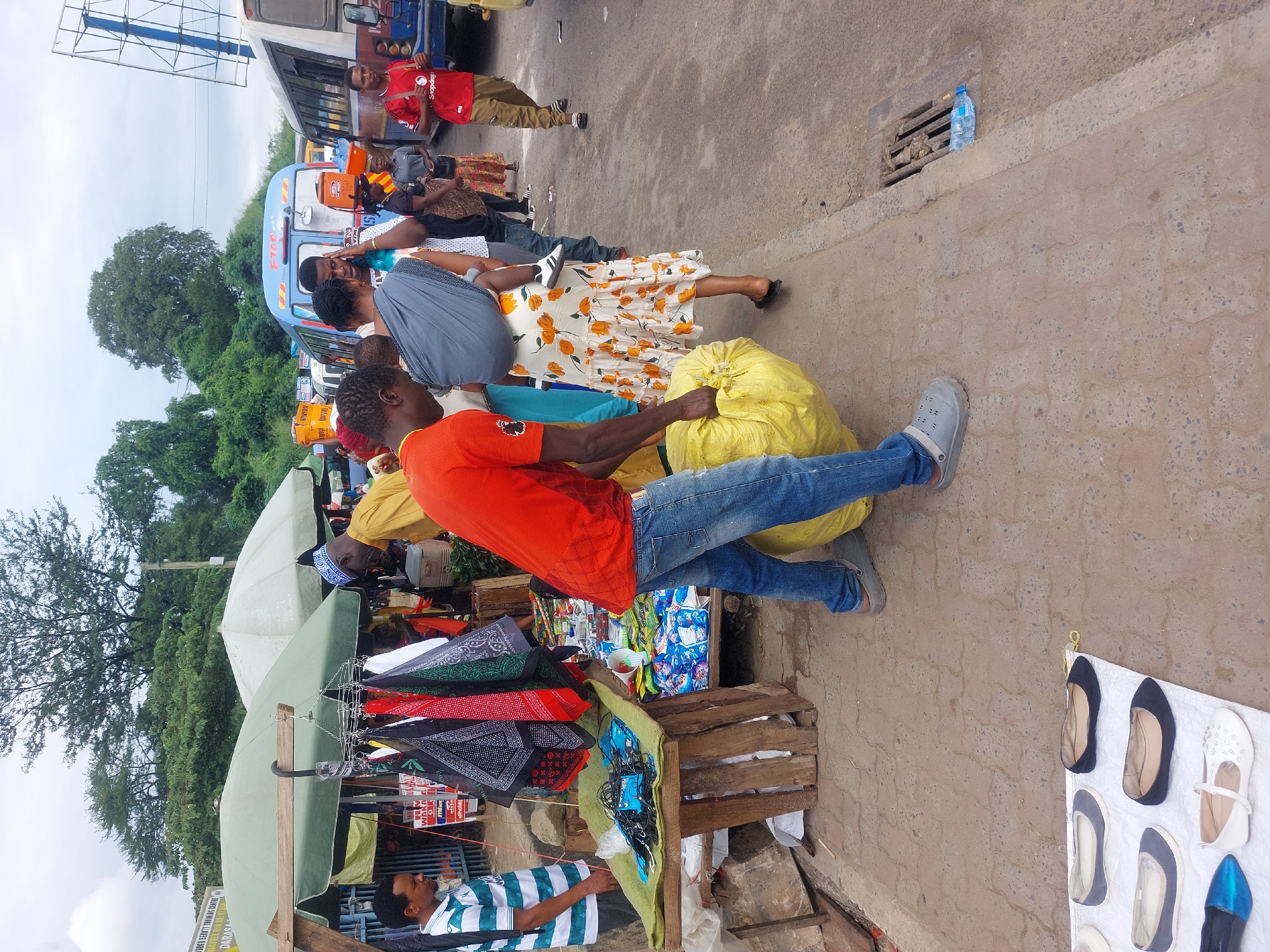 The image depicts a bustling street scene in Dar es-Salaam, Tanzania. People are waiting near a bus. A man in an orange shirt and jeans carries a large yellow sack. Adjacent to him, a vendor's stall is set up, displaying bandanas, small snack bags, and other items, under an umbrella. Footwear is displayed on the ground. A person in a striped shirt is at the front of the stall. In the background, there are trees and buildings.