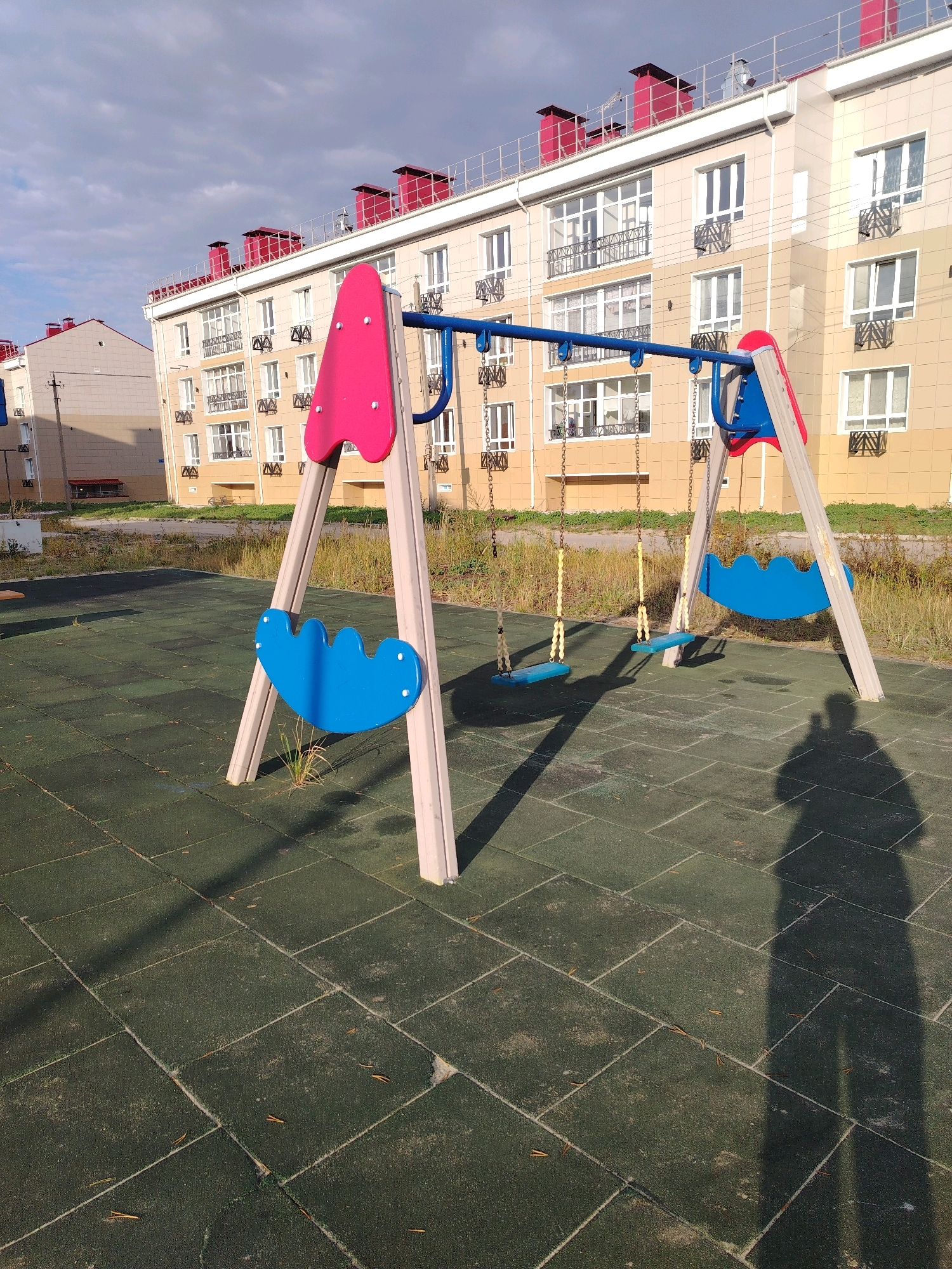 In Muravlenko, Russia, a playground features a bright pink and blue swing set with two blue seats. The swings are suspended from a blue metal bar supported by beige A-frame structures. The playground surface is made of dark green rubber tiles. In the background, a modern, multi-story apartment building with light-colored facade and red-trimmed roofs is visible. The sky is partly cloudy with a mix of blue sky and grey clouds. A shadow of a person is cast on the playground floor, indicating the presence of an observer or photographer. The scene is calm and still, suggesting an unoccupied public space.