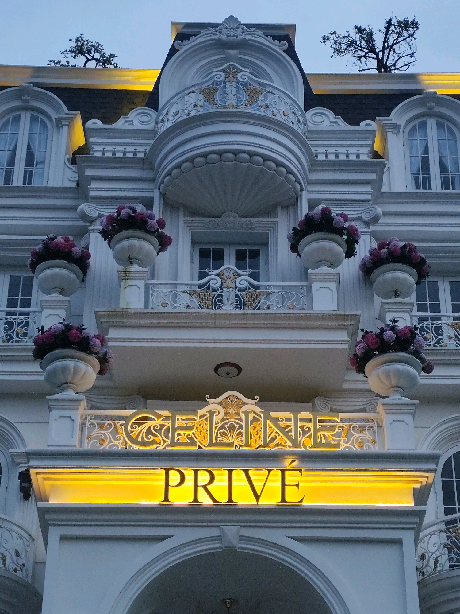 The image captures a detailed exterior view of a building in Lâm Viên - Đà Lạt, Vietnam, with a distinct architectural style. The building is predominantly white, featuring ornate decorative elements like moldings, columns, and balconies with intricate wrought-iron railings. Several large, decorative planters filled with pink and purple flowers are strategically placed on ledges. Above the entrance, a prominent illuminated sign displays the words "CÉLINE PRIVE" in gold lettering against a bright yellow background, suggesting a high-end establishment. Arched doorways and windows with intricate framing are visible. The sky above is overcast and pale, indicating a possibly twilight or cloudy day. Small trees with bare branches are visible on the rooftop, adding a touch of nature to the urban facade. No people are visible in the frame.