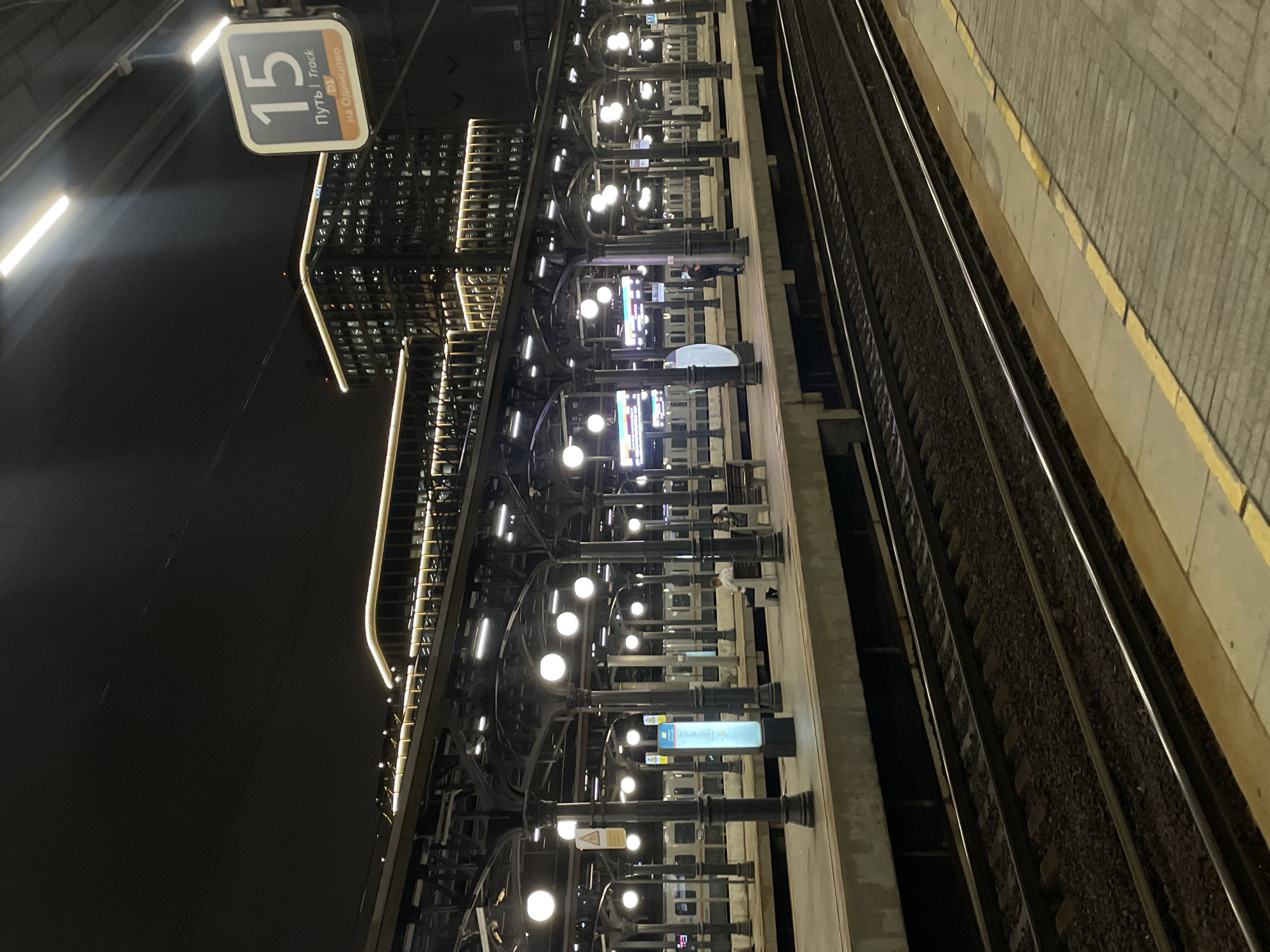 At night, on a train platform in Moscow, Russia, a sign labeled "15" indicates Track 15, which leads to Odintsovo. The platform is illuminated by spherical lights mounted on ornate, dark-colored columns that support a covered roof structure. In the background, a modern building with illuminated windows and decorative lighting stands out against the dark sky. Several trains are visible on the tracks. A few people are present on the platform; one person sits on a bench, and another pair stands near a pillar. The train tracks are clearly visible in the foreground, with gravel ballast between the rails. The platform surface is paved and marked with yellow lines along the edge.
