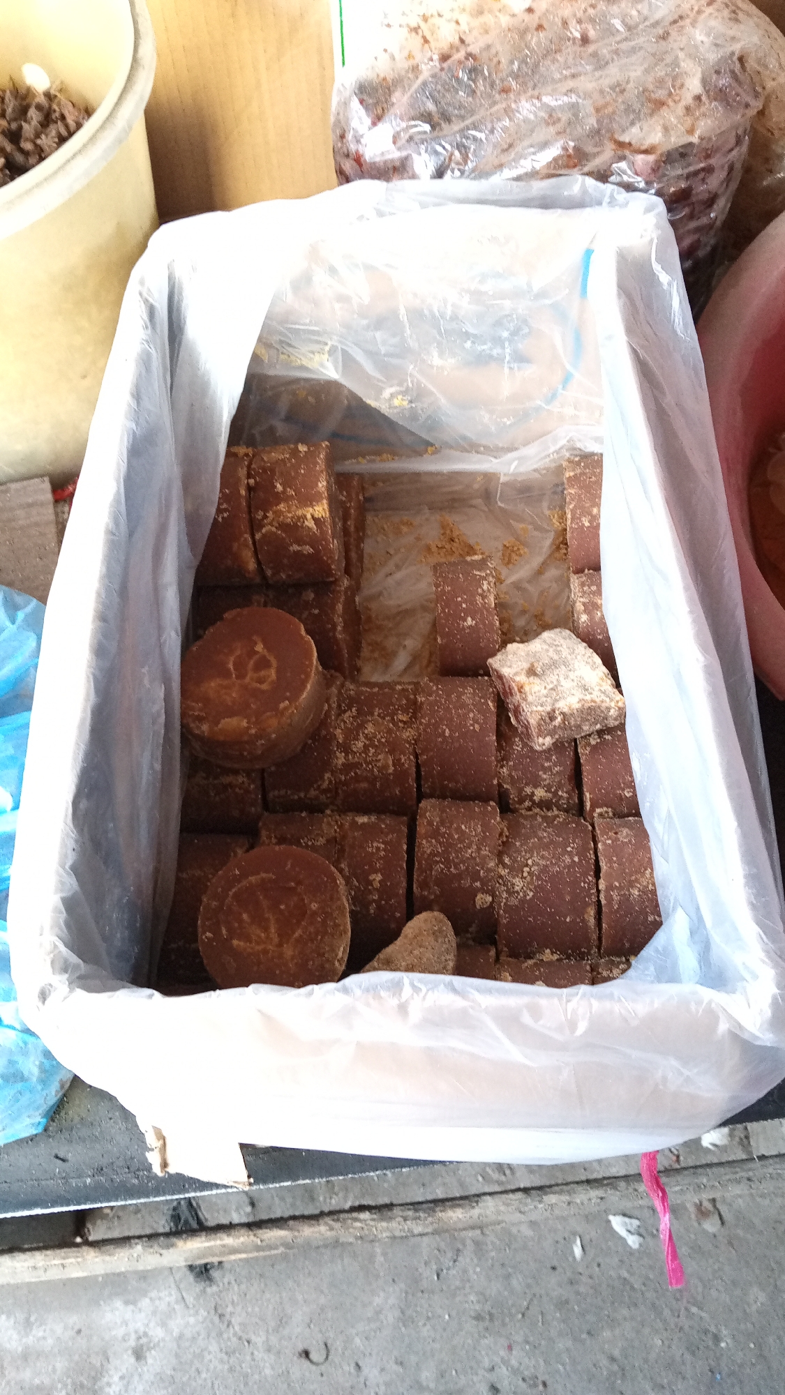 The image displays a white plastic-lined cardboard box filled with numerous blocks of brown sugar. The sugar blocks are of two shapes: cylindrical and rectangular prisms. Some of the blocks are covered in granulated sugar. In the background, to the left, is a portion of a light-colored plastic bucket containing a dark granular substance. To the upper right, a large plastic bag holds a dark, possibly sticky, material. The scene appears to be an outdoor market stall in Puloara, Indonesia, suggested by the rustic wooden surface beneath the box and the overall presentation of bulk goods.