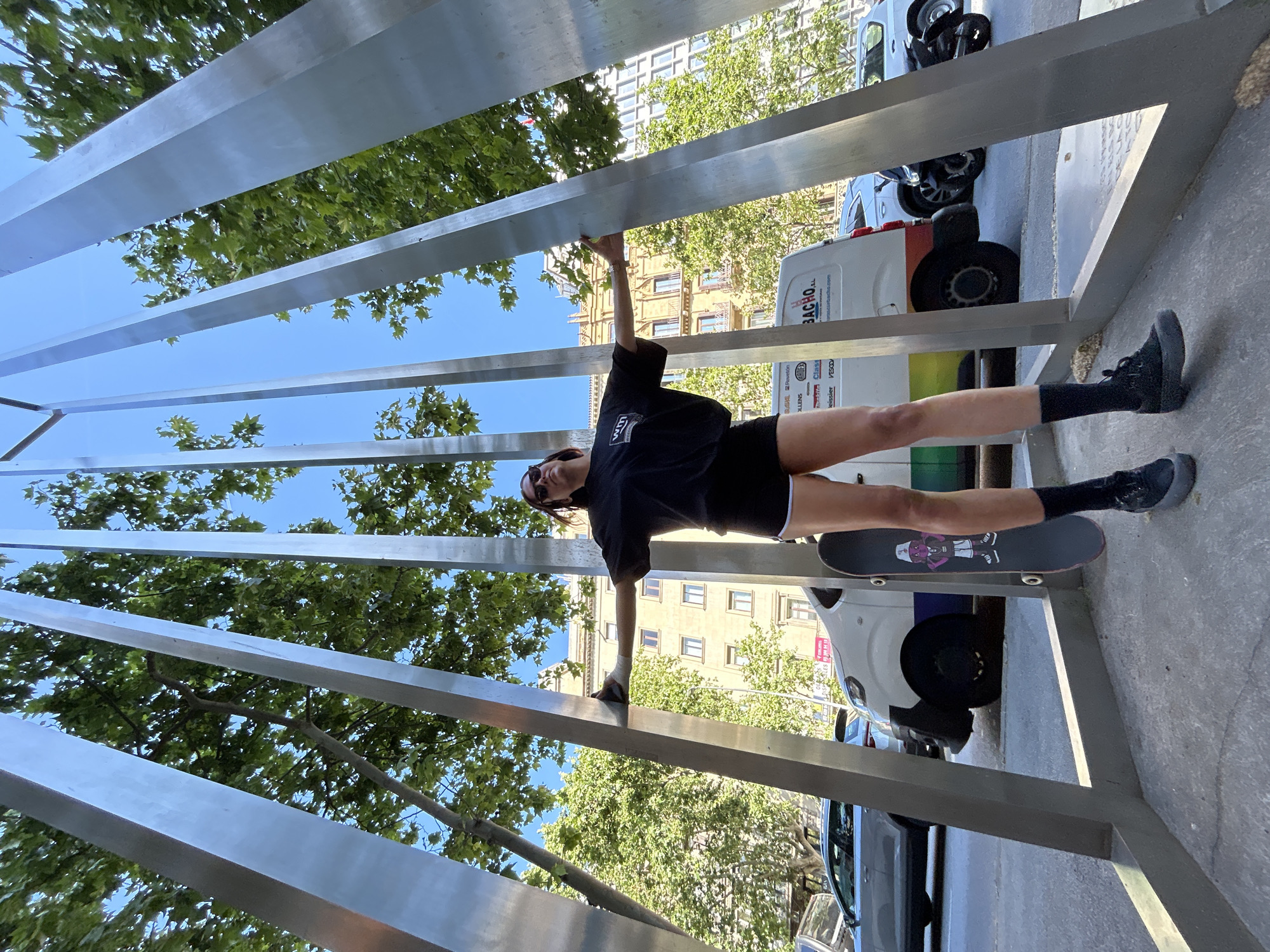 A person, seen from below, is walking with their hands and feet on a metal art structure. The person is wearing a black t-shirt, black shorts, black socks, and black sneakers. The person's feet are on a skateboard as the person walks across the art structure. The structure has parallel metal beams that allow views of buildings, trees, parked vehicles, and blue sky. The van visible behind the person has the name "BACHO" on the side. The trees and buildings are consistent with an urban environment in Barcelona, Spain.