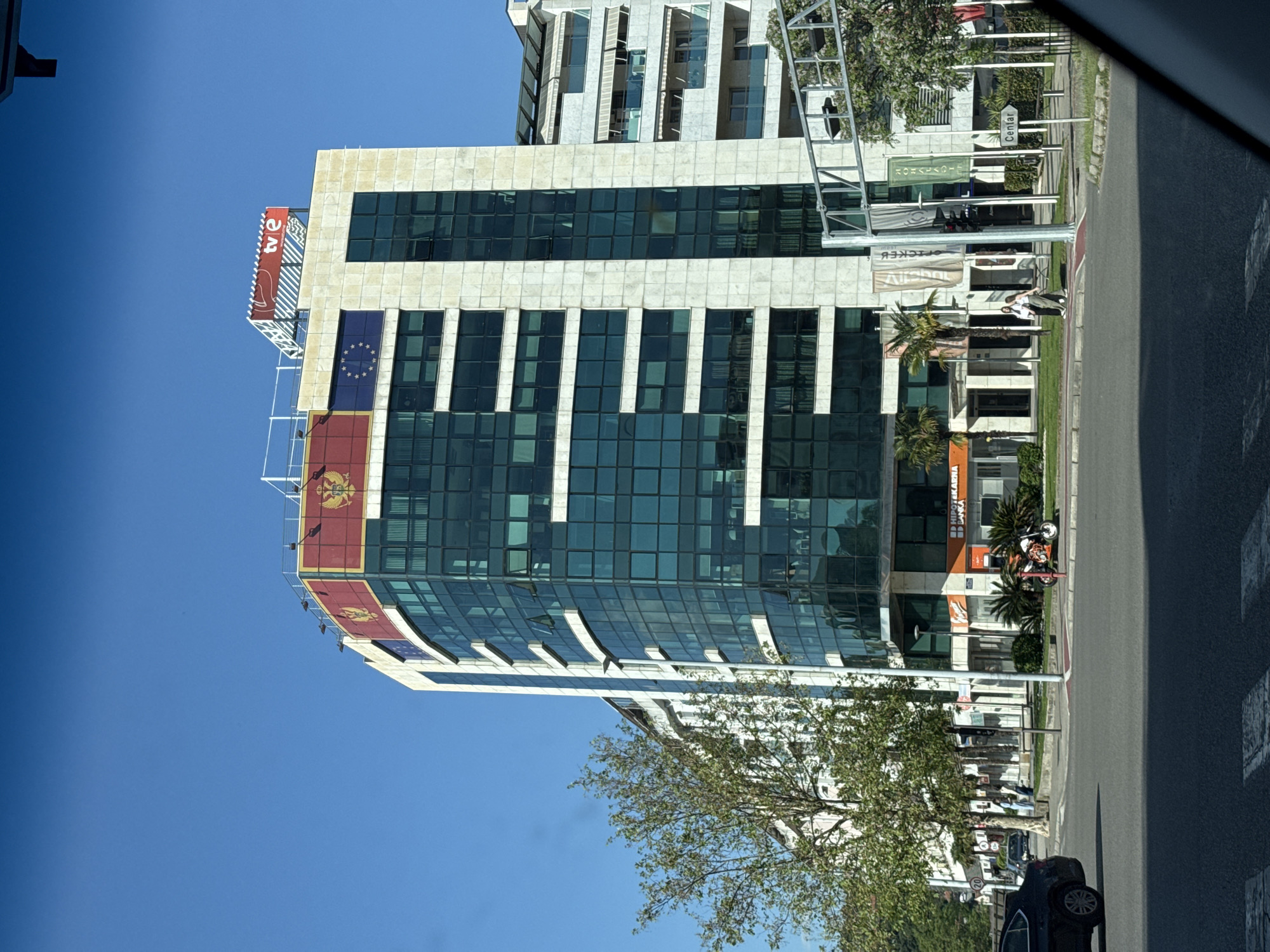 The image captures a modern building with a glass facade in an urban environment. The building is adorned with a "TVE" logo and what appears to be flags, possibly representing the European Union and a country. The scene is framed by a clear blue sky above and street-level details below, including trees, sidewalks, and a road. There are also street signs visible, and what appears to be a person seated on a bench by the sidewalk. A parked car is visible in the lower right corner. The location is an unknown city in an unknown country.