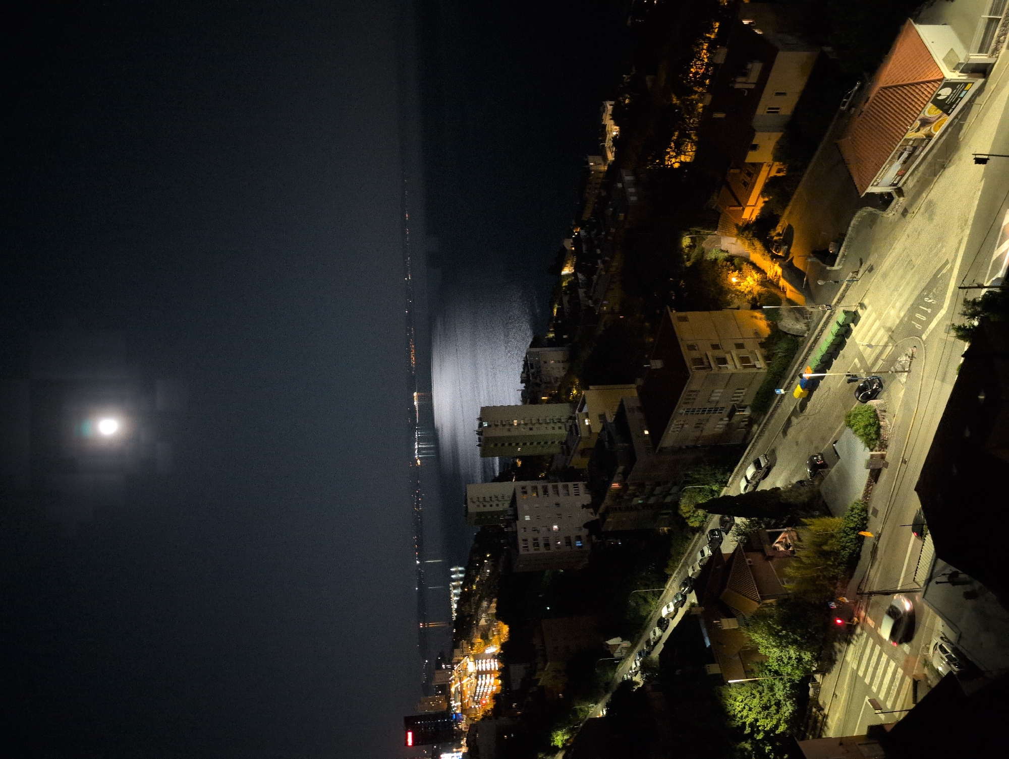 This is a night-time aerial view of a coastal city, likely in Rijeka, Croatia, with the full moon visible in the upper center of the frame. The scene is dominated by dark hues with artificial lights illuminating buildings and streets.
In the foreground, a street intersection is visible with crosswalks and streetlights. Several cars are parked along the side of the road, and at least two vehicles are in motion. One silver car is crossing the intersection from left to right, its headlights on, and a blurred white car is also visible on the street. Yellow and blue bins are lined up along the side of the road. A building with a red-tiled roof and a large banner is situated on the right side of the intersection.
Beyond the intersection, a densely populated urban area unfolds with apartment buildings of varying heights. Some buildings are illuminated from within, casting a warm glow, while others appear dark. Trees and vegetation are interspersed between the structures, adding a touch of green to the otherwise monochromatic night scene.
Further in the distance, a body of water, presumably the sea, stretches across the horizon. The moon's reflection shimmers on the water's surface, creating a luminous pathway. On the opposite shore, a line of city lights indicates further development and activity. A tall, dark building with illuminated windows stands out on the left side of the cityscape, near the waterfront. The overall impression is one of a lively city at night, with the serene presence of the sea and the moon overhead.