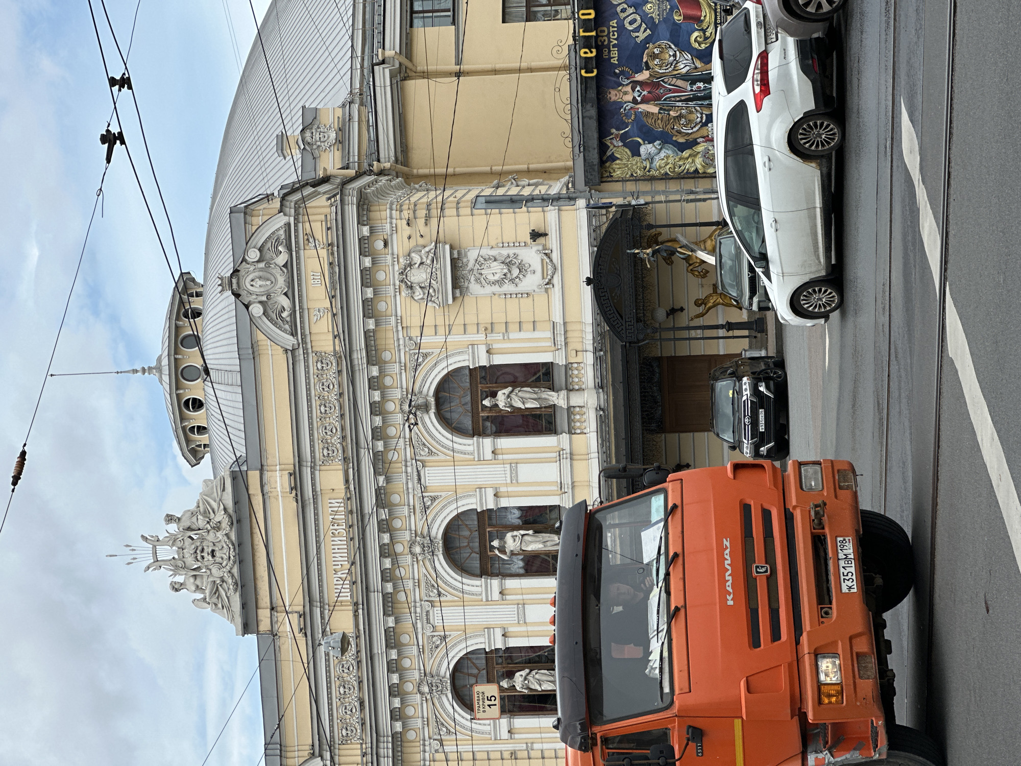 This is a medium-shot, eye-level, outdoor, daytime photograph of the historic Nikulin Circus on Tsvetnoy Boulevard in Moscow, Russia, although incorrectly identified as Saint Petersburg. In the foreground, a large, orange Kamaz truck is partially visible, facing left. The truck has the "KAMAZ" logo on its grille and a license plate reading "K351BM 198". To the right of the truck, a white Ford Focus station wagon is parked. Behind these vehicles, the ornate yellow facade of the circus building is prominent. The building features classical architectural elements, including statues in niches, decorative reliefs, and arched windows. The circus's name, "ЦИРК ЧИНИЗЕЛЛИ" (Circus Ciniselli), is visible in large letters on the facade. A large, colorful poster depicting performers and tigers is displayed on the right side of the building. Overhead power lines crisscross the sky, which is overcast. The overall scene depicts a busy urban environment with a mix of vehicles and historical architecture.