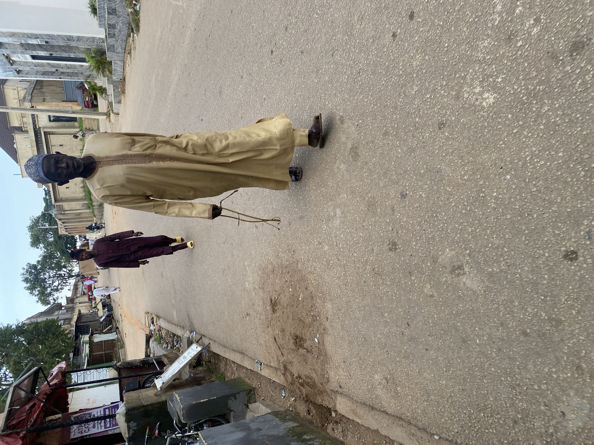 The image depicts a street scene in Jos, Nigeria. In the foreground, an elderly man, wearing a traditional light brown kaftan and a patterned cap, walks towards the camera, holding a string of dark prayer beads. He is wearing dark sandals. Behind him, another man in a dark maroon outfit and yellow sandals walks away from the camera. The street is paved with asphalt, with debris and loose dirt along the sides. Buildings line the street, with some appearing to be shops or commercial establishments. The sky is overcast.