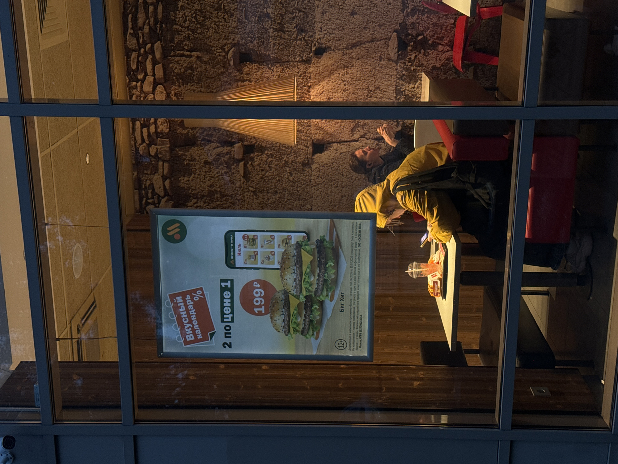 This image captures an evening scene through the window of a fast-food establishment, likely in Saint Petersburg, Russia, given the Cyrillic text on the advertisement. Inside, two individuals are seated at a table. One person, wearing a yellow hooded jacket and a backpack, is focused on their phone, with a drink and food items on the table. Another individual, partially obscured, is seated opposite them, also looking down.
The restaurant's interior features a rustic stone wall and warm lighting from stylized lamps. A prominent advertisement poster displays images of hamburgers and promotional text in Russian, indicating a "Tasty Calendar" and a "2 for 1" offer on a "Big Hit" burger, priced at 199 rubles. The poster also includes a 12+ age rating.
The exterior view is framed by the building's modern facade and window panes, reflecting some of the interior light and the dim exterior environment. The overall impression is one of a casual dining experience on a cool evening.