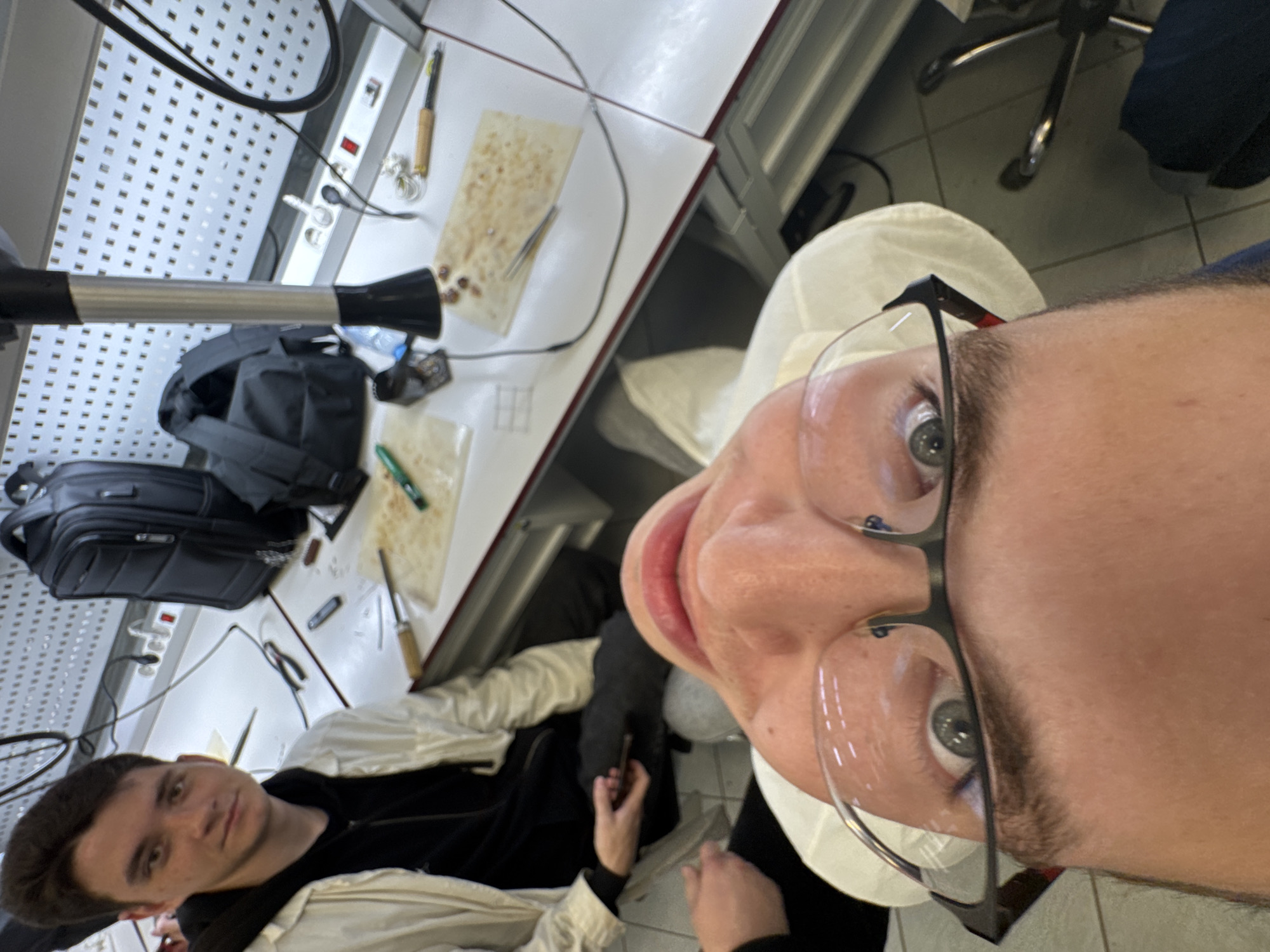 This image captures an indoor scene, likely a workshop or lab, from a rotated, close-up, and slightly distorted perspective, as if taken from a low angle during a selfie.
In the immediate foreground, significantly magnified due to the close proximity, is the face of a male individual. He has fair skin, light-colored eyes (appearing gray or green), and is wearing clear-framed glasses with dark temples. Only his eyes, nose, and mouth are clearly visible, with a small portion of his forehead and chin, and short dark hair around his temples. He has a neutral expression, looking directly towards the camera. A white collar, possibly from a lab coat or protective garment, is visible near his chin.
To the left and slightly further back in the frame, another young male is seated. He has fair skin and dark hair, looking calmly towards the camera. He is wearing a dark top, possibly a sweater or hoodie, covered by a light-colored (white or cream) jacket. One of his hands is visible, appearing to hold a dark object, possibly a mobile phone.
The setting is a workbench or lab table with a light-colored surface, featuring a pattern of small holes, possibly indicating a pegboard or specialized work surface. On the table, various tools and items are scattered. These include a black backpack, a power strip with a red switch and outlets, wires and cables, and what appears to be a small black fan or blower. Several small, reddish-brown, irregularly shaped objects (possibly beads, stones, or components) are laid out on light-colored, textured mats or cloths. Other tools like tweezers and a pen-like instrument with a wooden handle are also visible. A clear plastic bottle is partially seen. Behind the seated person, part of an office chair with chrome legs is visible.
The overall scene suggests an environment dedicated to intricate work, possibly involving crafting, jewelry making, or electronics. The presence of two individuals suggests collaboration or shared workspace. No discernible text is visible in the image. The location context provided is Perm, Russia.