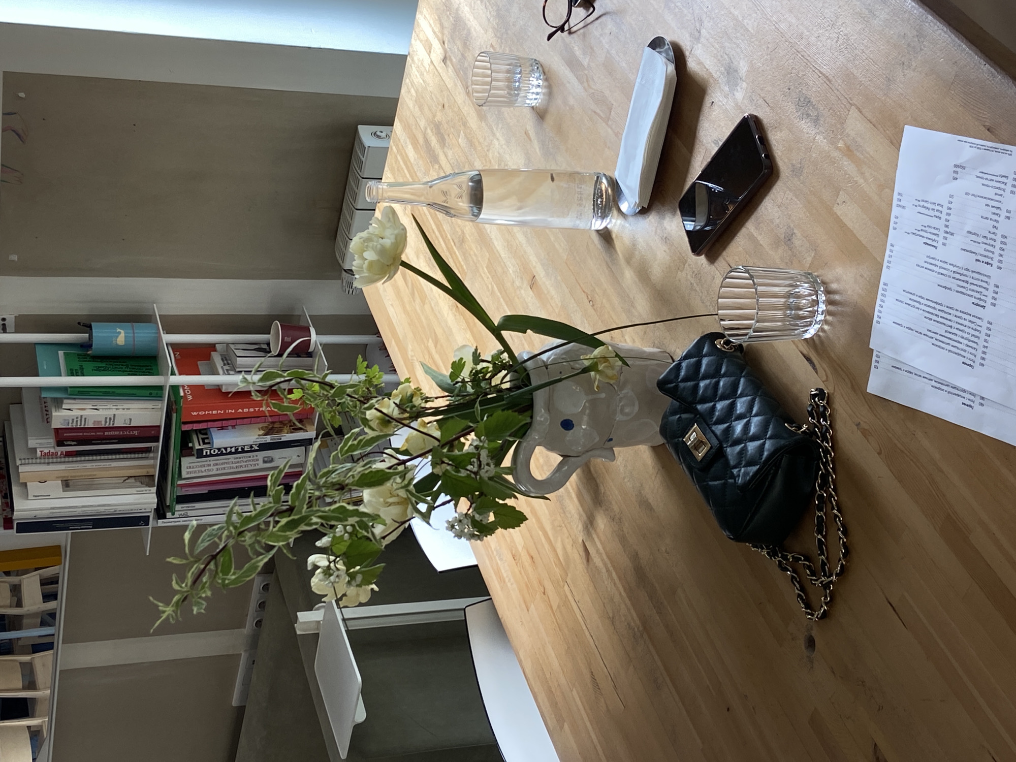 The image presents a tabletop scene, likely in an indoor setting, dominated by a mix of personal items and dining-related objects. On the table, there is a glass bottle, partially filled with a transparent liquid, possibly water, along with a drinking glass. A cell phone rests nearby. Additionally, there are some pieces of paper, possibly menus or notes, spread out on the surface. A small black quilted purse with a gold chain is also present.
A vase containing flowers adds a natural element to the composition. A shelf of books and other items can be seen behind the table and vase.
Given the inclusion of a Russian menu and book titles in Russian, the scene is likely located in Russia.