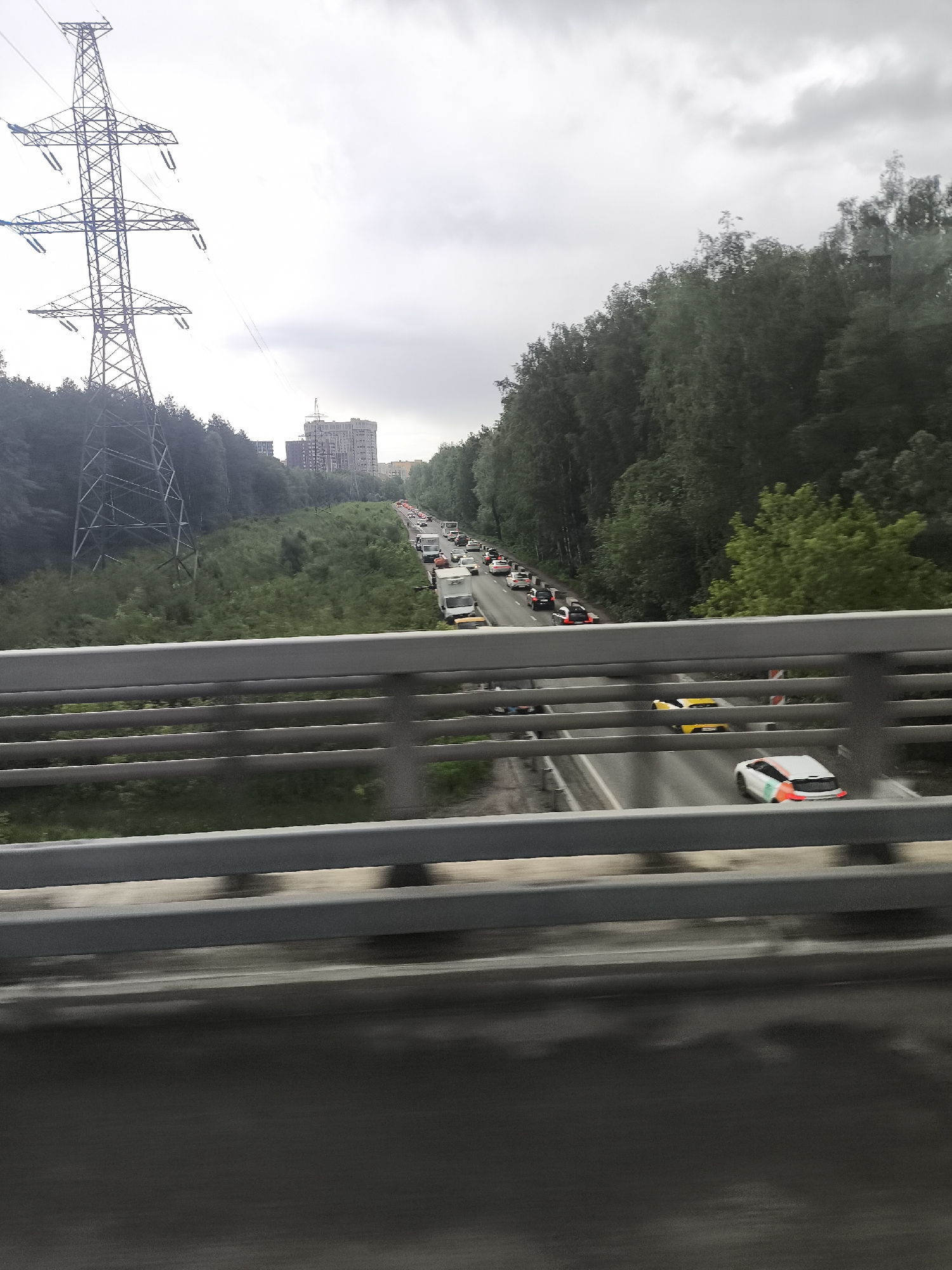 The image shows a highway scene in the city of Unknown, Unknown. The view is taken from a bridge, overlooking a multi-lane road filled with traffic. A convoy of vehicles, including cars and a utility vehicle, are moving slowly in the distance. The roadside is lined with dense green trees. A tall, gray power transmission tower is visible to the left. In the background, a multi-story building is seen through the trees. The sky is overcast with gray clouds. The bridge's gray metal railing is in the foreground.