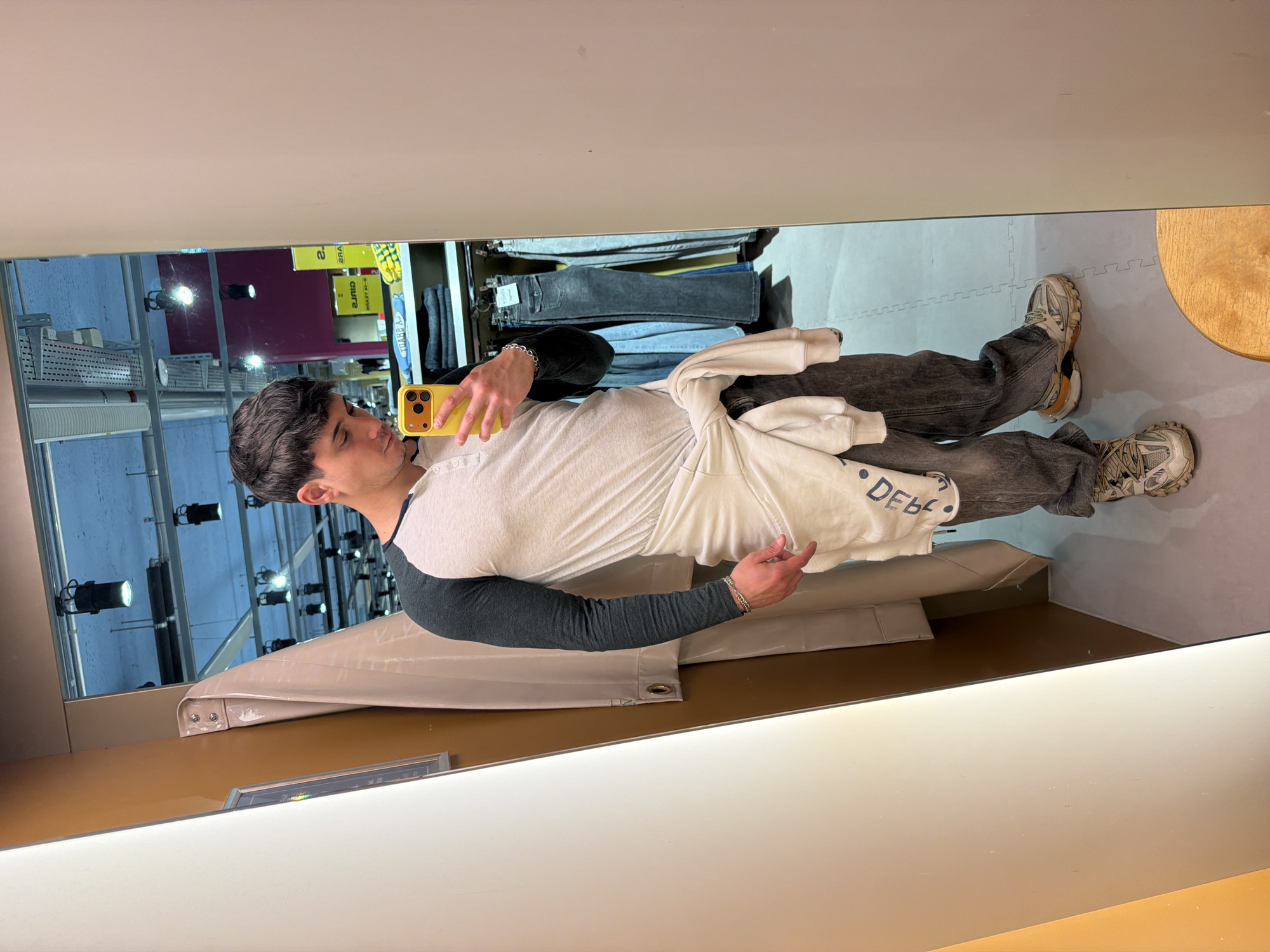 A man stands in a mirrored room, likely a clothing store fitting room. He is taking a selfie with a yellow smartphone. He is wearing a white and grey raglan shirt, dark grey jeans, and sneakers. He has a white sweatshirt tied around his waist. Garments hang on racks in the background. The room appears to be in a retail environment, possibly in Spreitenbach, Switzerland, given the presence of retail shelving, lighting, and mirrored walls typically found in clothing stores.