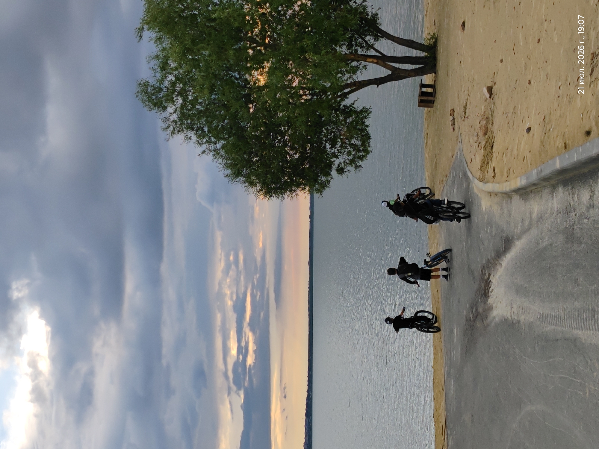 The image depicts a group of three individuals on bicycles near a body of water, likely a lake, on a cloudy day. The individuals are positioned on a sandy shore adjacent to a paved area. One person is riding a bicycle forward on the paved path, while another is stationary on a bicycle on the sand. A third person is also on a bicycle on the sand, facing away from the camera. A large tree with green foliage stands on the right side of the image, near the water's edge. The sky is overcast with dramatic cloud formations, suggesting either an approaching storm or the aftermath of one. The water appears calm. In the background, a distant shoreline with buildings can be seen. The bottom right corner of the image contains a timestamp in Cyrillic script: "21 июл. 2026 г., 19:07," which translates to "July 21, 2026, 19:07." This timestamp, combined with the location context of Kokshetau, Kazakhstan, places the scene as an outdoor activity by a lake in Kokshetau on the specified date and time.