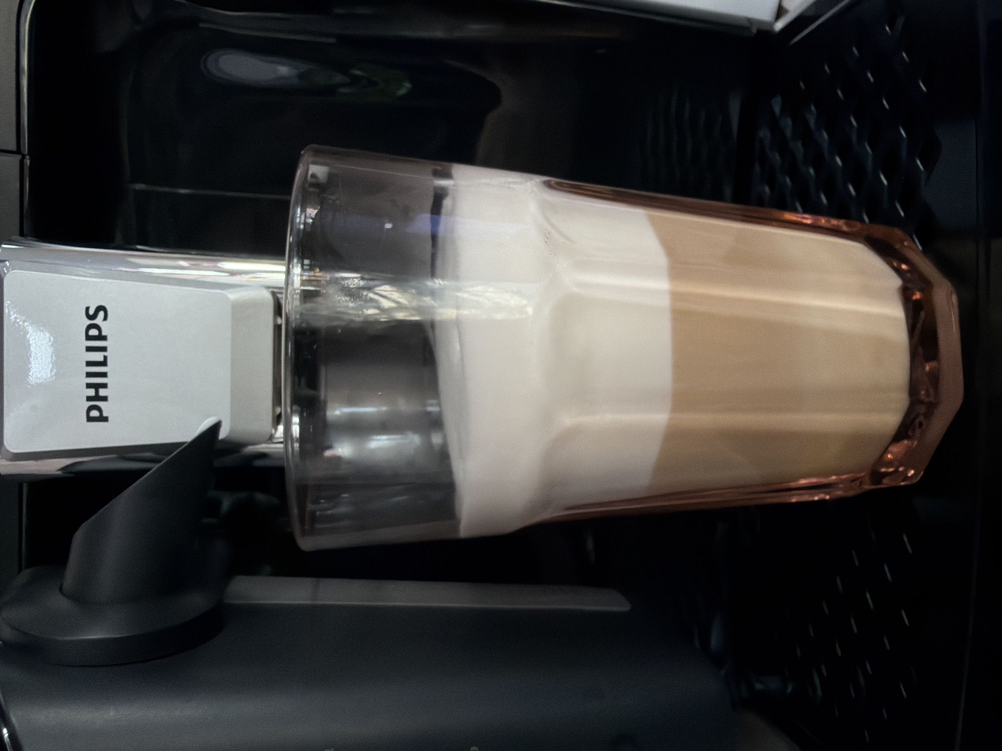An overhead shot shows a glass of latte resting on a coffee machine. The glass is full, with distinct layers of milk foam and coffee. The coffee machine is black with a white panel that displays the Philips logo. The background is dark and out of focus. The scene is indoors, likely a kitchen or cafe.