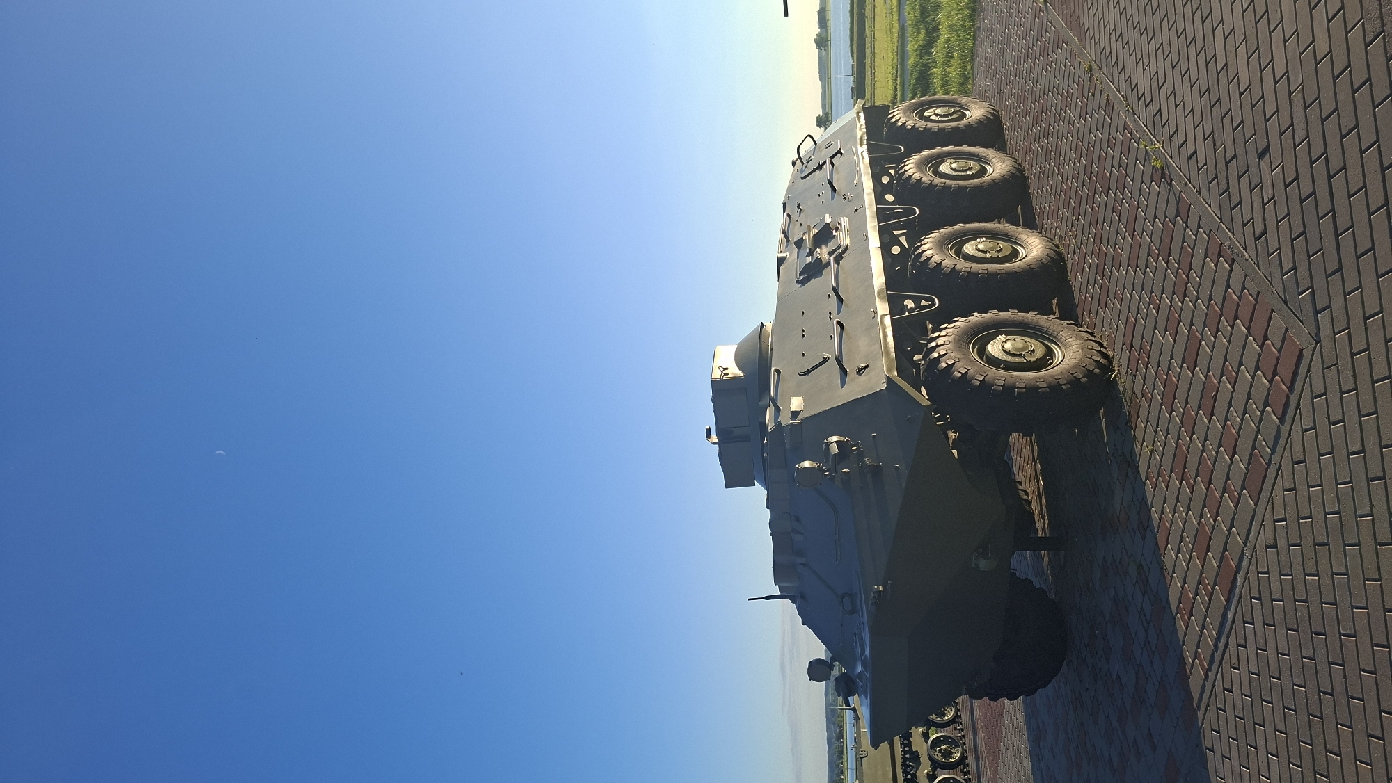 An eight-wheeled armored personnel carrier is parked on a paved area under a clear blue sky with a faint crescent moon visible. The vehicle is dark green and has a multi-faceted hull. It is positioned facing slightly left, with its front angled towards the viewer. The paved area is composed of rectangular bricks in shades of gray and red. Beyond the paved area, there is green grass and a body of water with trees in the distance. The scene appears to be outdoors, possibly at a museum or memorial in the city of Smalyavichy, Belarus. No people are present in the image.