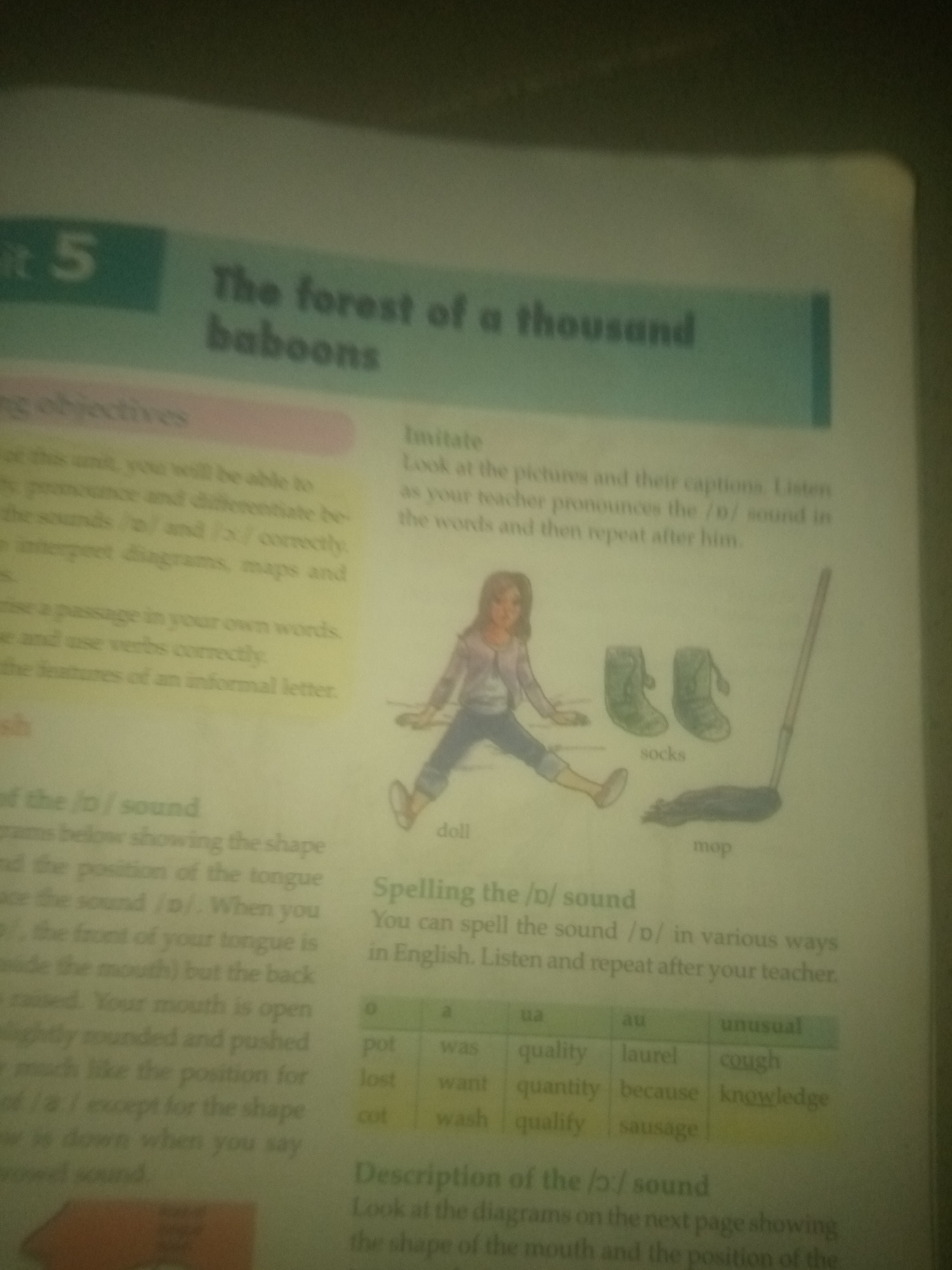The image displays a page from a book, likely an educational text. The page is divided into sections with text and illustrations.
On the left side of the page, there is a heading "it 5" and below it, "The forest of a thousand baboons." Further down, there is a section titled "ng objectives" followed by bullet points detailing learning goals.
On the right side, there is a section titled "Imitate" with instructions to look at pictures and repeat words. Below this, there is an illustration of a girl sitting on the floor with several objects: a doll, two pairs of boots labeled "socks," and a mop.
Below the illustration, there is a section titled "Spelling the /ɔ/ sound" with a table containing words organized under different sound categories.
Further down the page, there is a section titled "Description of the /ɔ/ sound" with instructions to look at diagrams.
The image is brightly lit, suggesting it was taken indoors. The text is generally legible, though some parts are slightly blurred or out of focus. The page appears to be a typical classroom or textbook material, focusing on language and literacy skills, specifically pronunciation and spelling of the /ɔ/ sound. There is no information in the image that provides any context about the city of Jauro Yinu, Nigeria.