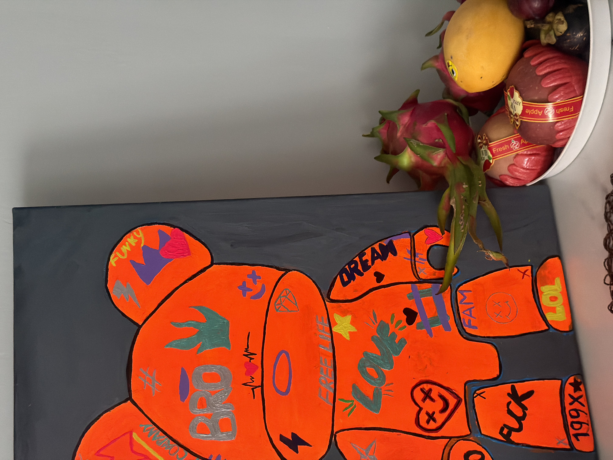 An oil painting of an orange bear with graffiti is displayed on a gray wall. The bear has "BRO" written on its head, "LOVE" on its chest, and "DREAM" on its arm. The painting is in the foreground, and a bowl of fruit, including dragon fruit, apples, a mango, and mangosteen, is to its right. The scene appears to be indoors, possibly in a home or gallery, with a plain gray wall in the background. There is no indication of the city of Nha Trang, Vietnam, or any other specific location.