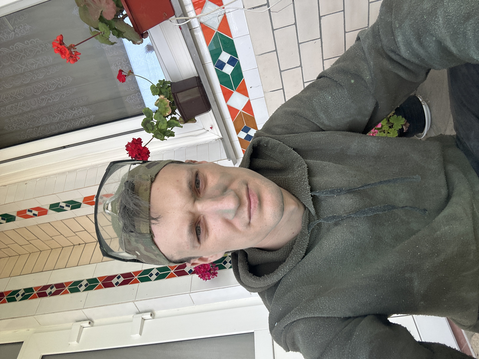A man wearing a camouflage baseball cap and safety glasses, with a green hooded sweatshirt and visible dust on it, poses for a selfie outdoors. He has some green dust or paint on his face. In the background, there are red flowers in pots, a window with a decorative curtain, and a wall with colorful tile accents. The wall is made of light-colored bricks. The overall scene suggests an outdoor setting, possibly a residential building's porch or entrance. The location is identified as Makiv, Ukraine.