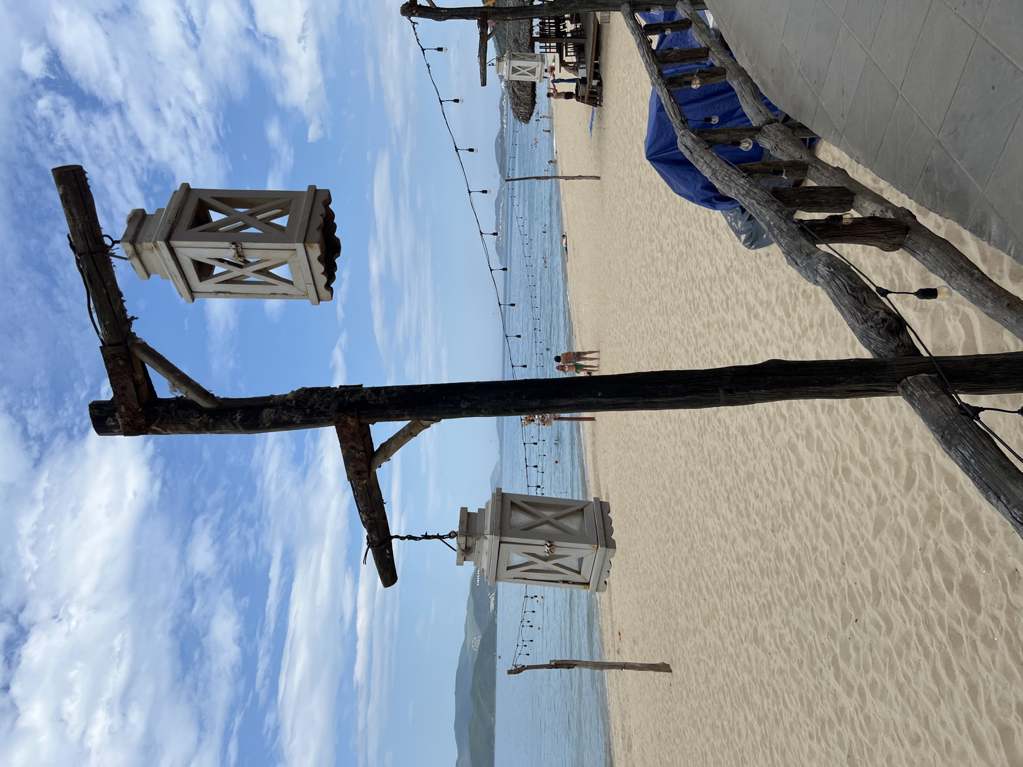 This is a photograph of a beach in Nha Trang, Vietnam, likely taken in the afternoon. The foreground is dominated by a rustic wooden structure with two hanging lanterns, one in the upper right and another in the middle left of the frame. The lanterns are octagonal and appear to be made of light-colored wood. The wooden structure itself is made of rough, weathered logs, with string lights attached to it.
The beach is sandy and appears to be relatively clean. In the middle ground, a calm, blue sea stretches to the horizon, with a hazy mountain range in the distance. There are a few people visible on the beach and in the water. Some are swimming or wading, while others are walking or relaxing on the sand.
To the right of the frame, there is a thatched-roof structure, possibly a bar or restaurant, with tables and chairs. A blue tarp is visible behind the wooden railing in the foreground. The sky is partly cloudy with streaks of white clouds against a bright blue background. The overall impression is of a relaxed, tropical beach scene.