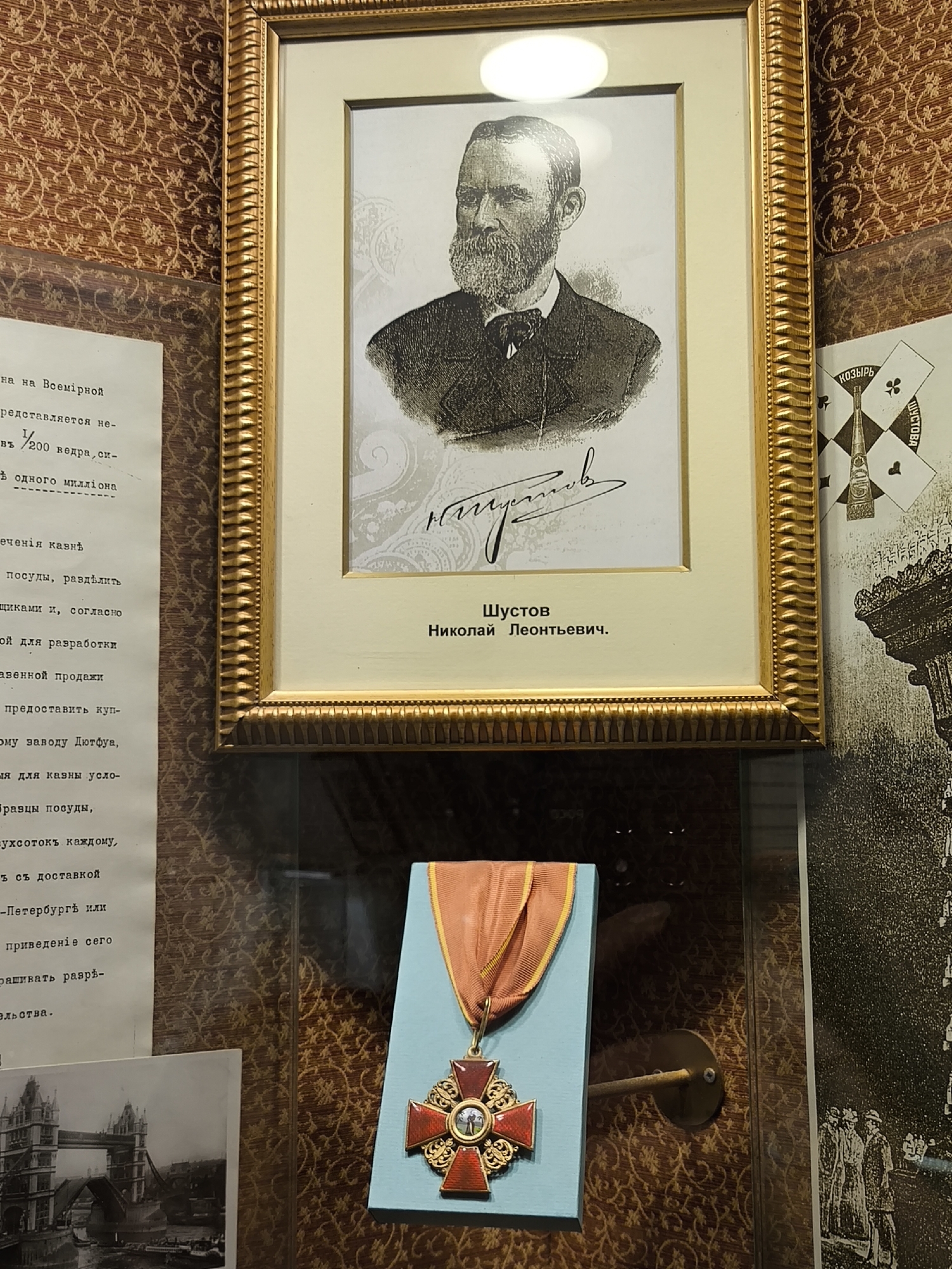 This media file shows a display of historical artifacts, likely in a museum setting in Saint Petersburg, Russia. The central subject is a framed portrait of a man with a beard, identified by the visible text as "Шустов Николай Леонтьевич" (Shustov Nikolai Leontievich). The portrait is a black and white etching or drawing, presented within an ornate golden frame.
To the left of the portrait, a document with text in Russian Cyrillic script is displayed. Some legible words include "Всемірной" (world), "редставляется" (is presented), "казнѣ" (treasury), and "заводу" (factory). There is also a mention of "Петербургѣ" (Petersburg).
Below the portrait, a medal on a stand is visible. The medal is red and gold, with a cross shape and a central circular element. It is suspended from a reddish-orange ribbon with a yellow border.
To the right of the portrait, there are some graphic elements that appear to be related to a brand or product, with the word "ШУСТОВА" (Shustova) visible. Further to the right, a darker, silhouetted image of buildings or people in a procession can be seen.
In the lower left corner, a black and white photograph shows a prominent bridge structure, likely the Tower Bridge in London, suggesting a broader historical context or connection beyond Saint Petersburg.
The overall setting appears to be an indoor exhibition, with patterned wallpaper on the walls and a glass display case protecting the artifacts. The lighting is even, suggesting an exhibition environment. There are no obvious indicators of time of day or weather. The emotion conveyed is one of historical reverence and preservation.