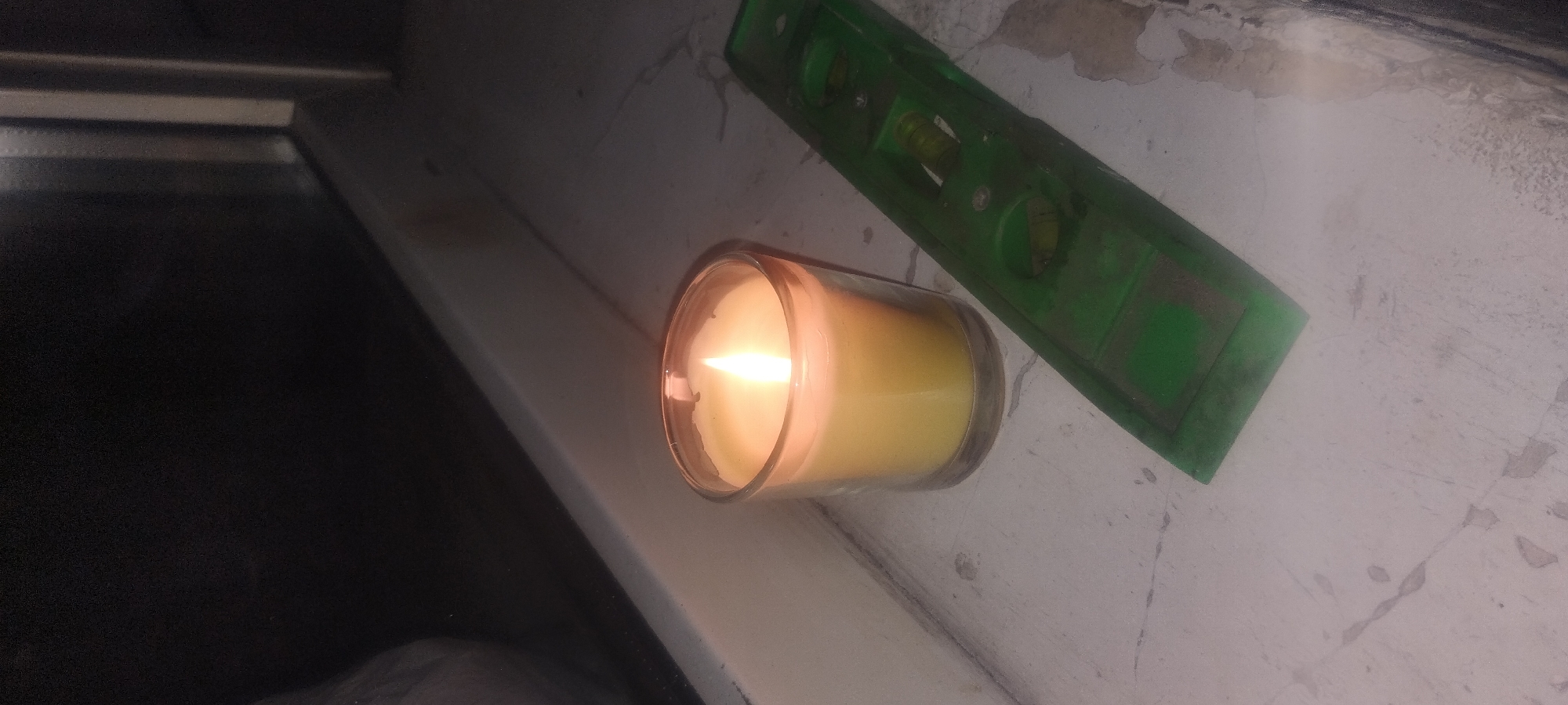 A lit yellow candle in a clear glass holder sits on a windowsill next to a green spirit level. The background is dark, suggesting it is nighttime or indoors with the lights off. The windowsill appears to be made of a rough, possibly painted, surface. The spirit level shows two bubble vials, indicating it is in use or has recently been used. The overall scene is dimly lit, with the candle flame being the primary source of light. There is no information in the image to determine the location in the city of Zaporizhzhia, Ukraine.