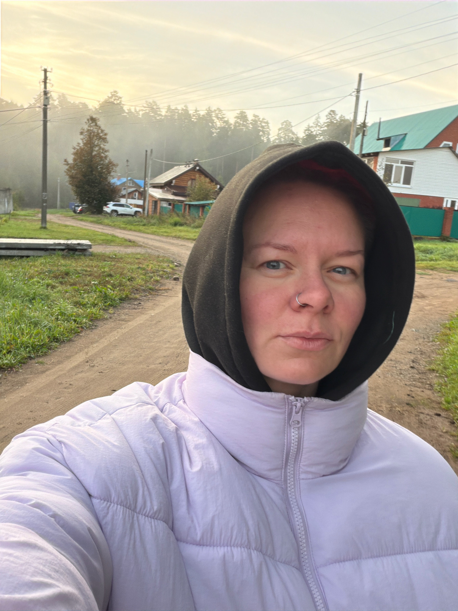 This is a selfie of a woman wearing a black hooded sweatshirt under a light purple puffer jacket. She has a nose ring and is standing on a dirt road in what appears to be a rural area. In the background, there are wooden houses, trees, power lines, and a white SUV. The sky is overcast and hazy, suggesting it might be early morning or a foggy day. The overall scene depicts a quiet, rustic setting. The location is confirmed to be the city of Chaikovsky, Russia, based on the provided context.