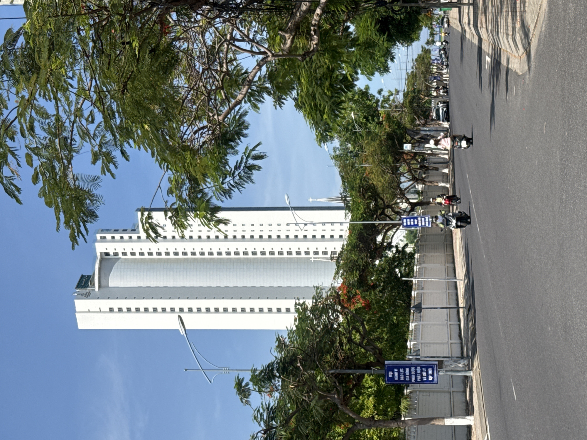 In the city of Bac Nha Trang Ward, Vietnam, a tall, white, modern high-rise building dominates the skyline under a clear blue sky. The building features a repeating pattern of dark rectangular windows. In the foreground, a paved road runs parallel to a white, decorative wall. Trees with lush green foliage and scattered red flowers line the street. Several motorcycles and at least two cars are visible on the road. One motorcycle rider, a person in a pink shirt, is facing away from the camera. Another rider in a white helmet is also visible. A blue sign with text in Vietnamese is posted on a pole next to the road. The overall scene depicts a sunny day in an urban environment with significant greenery.